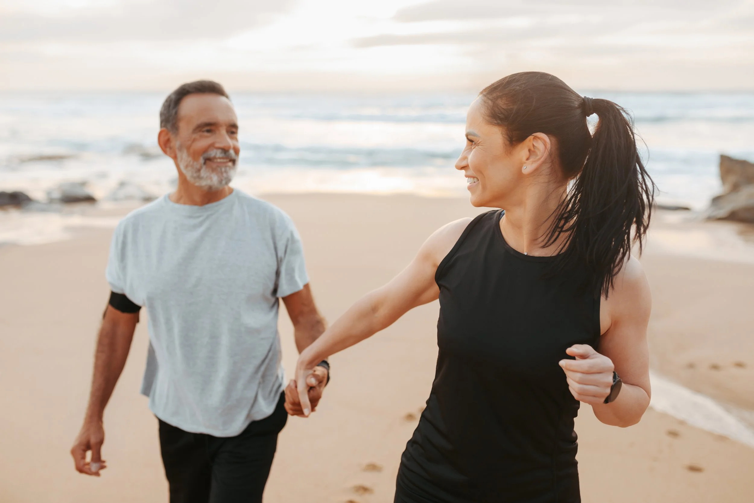 A smiling woman and man holding hands and running on the beach at sunset.
