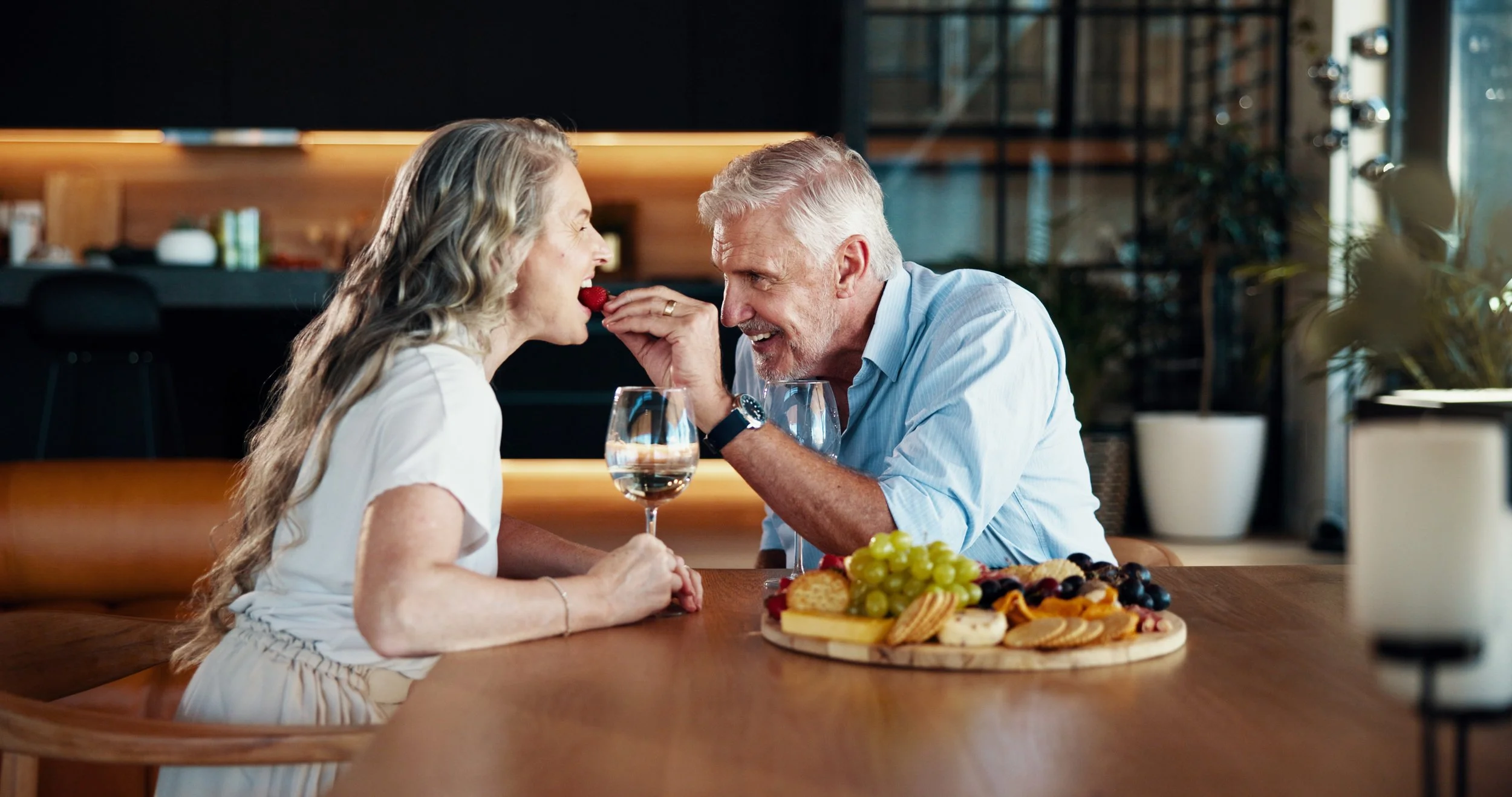 An elderly man and woman with gray hair smiling at each other at a dining table, with the woman feeding the man a strawberry, and a large platter of fruit and desserts on the table.