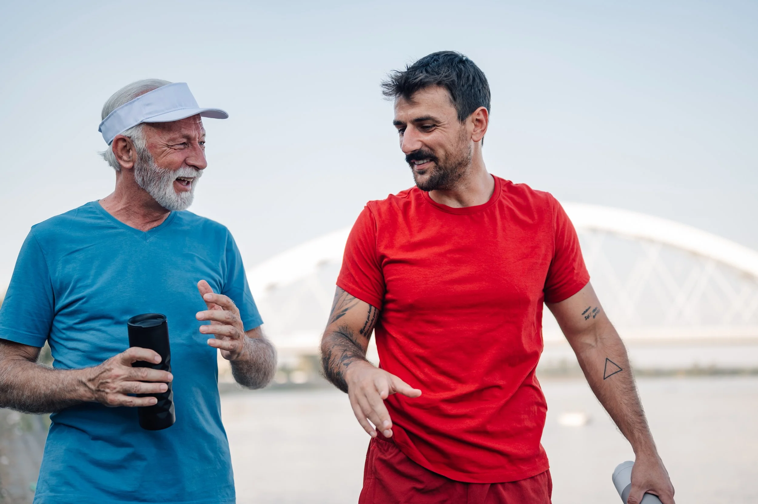 Two men, one older with a white beard and wearing a visor, and one younger with dark hair and tattoos, are smiling and talking outdoors during daytime.