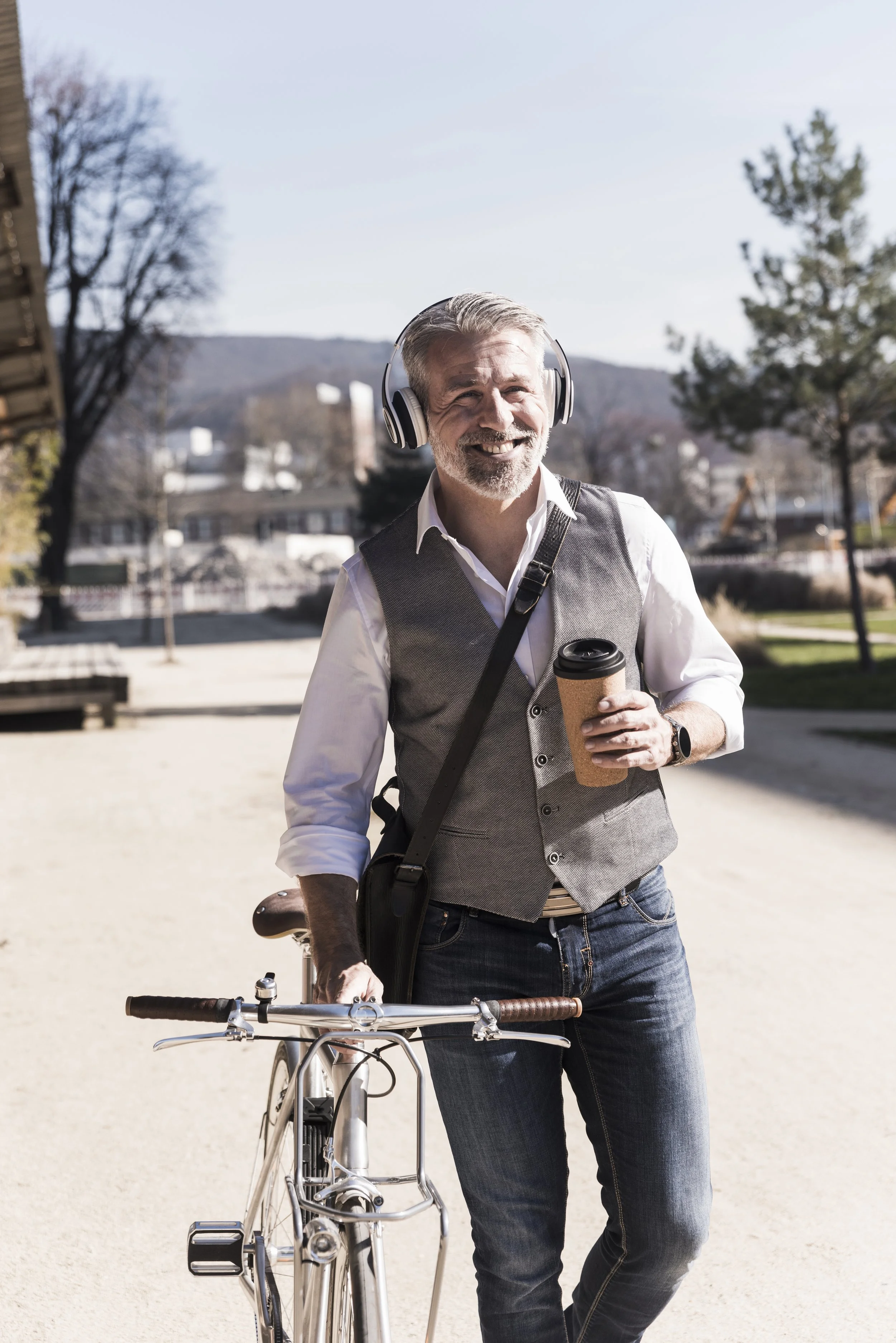 A man with gray hair and a beard, smiling, wearing headphones, a white shirt, a gray vest, and jeans, standing outdoors with a bicycle, holding a coffee cup, with trees and buildings in the background on a sunny day.
