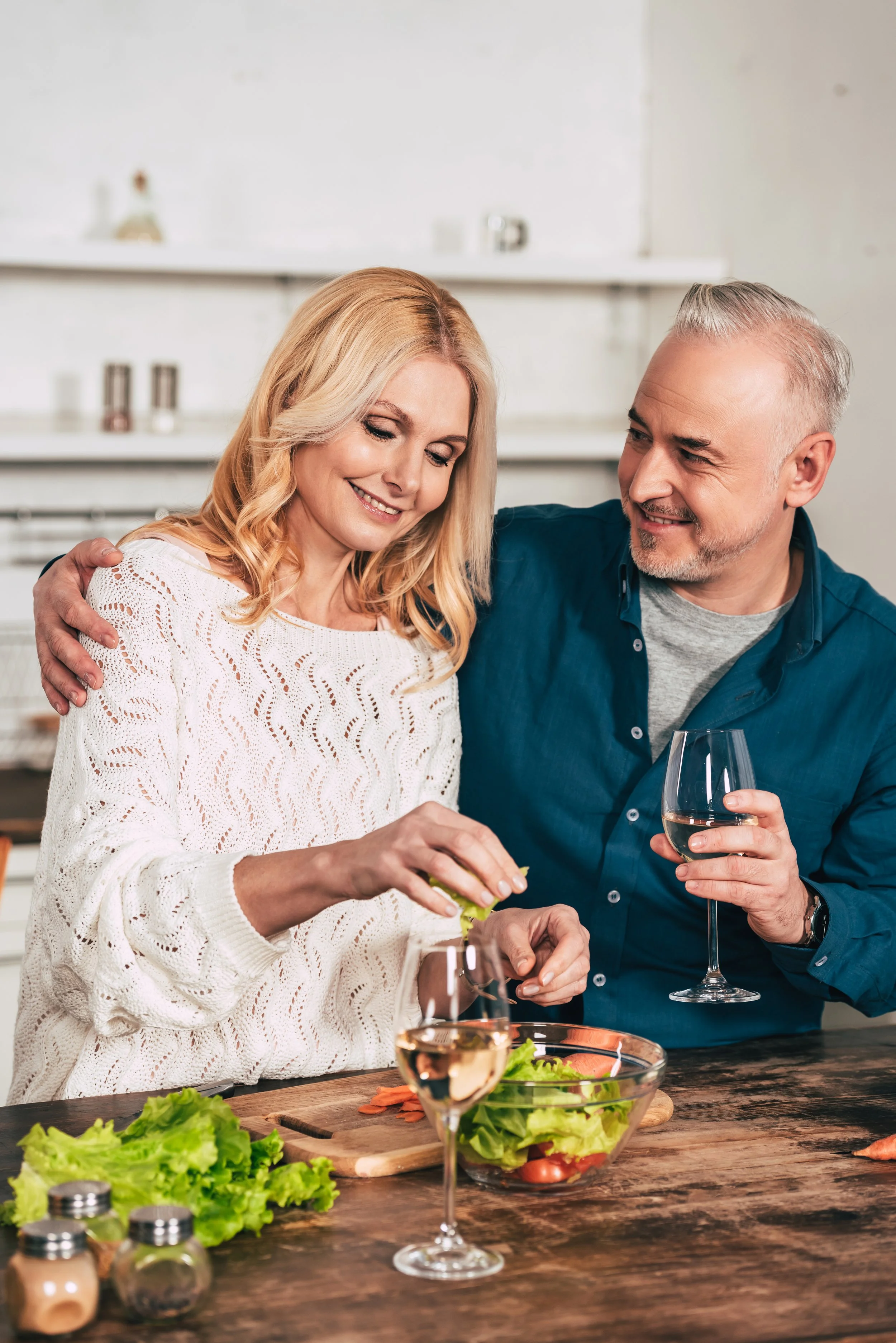 A middle-aged woman and man sharing a moment at a dinner table in a bright kitchen, with food and drinks including a glass of white wine, a salad, and fresh vegetables.