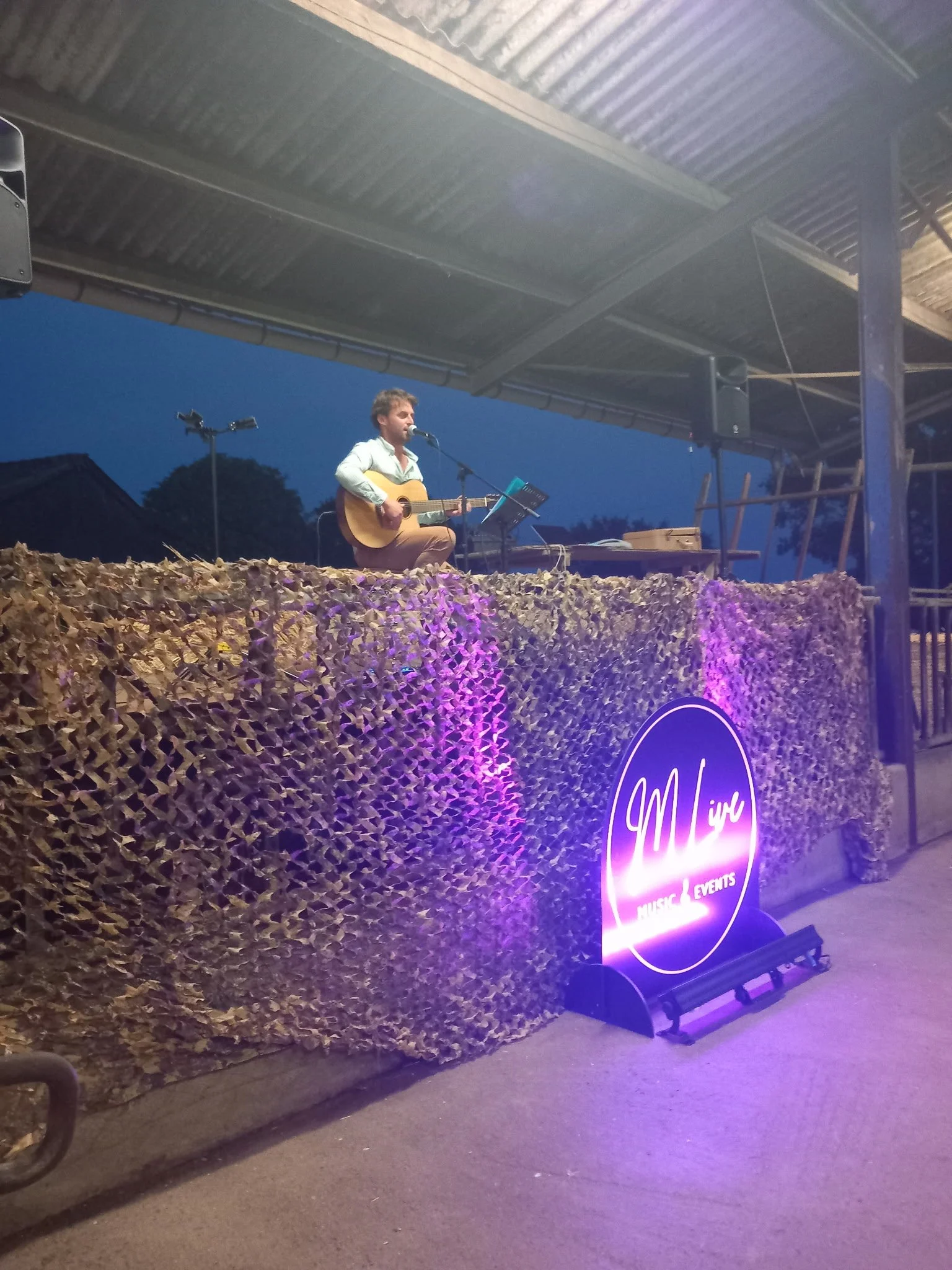 Scène nocturne avec un homme jouant de la guitare et chantant sur une scène en plein air, décorée avec un mur de feuillage et un panneau lumineux indiquant 'Music & Events'.