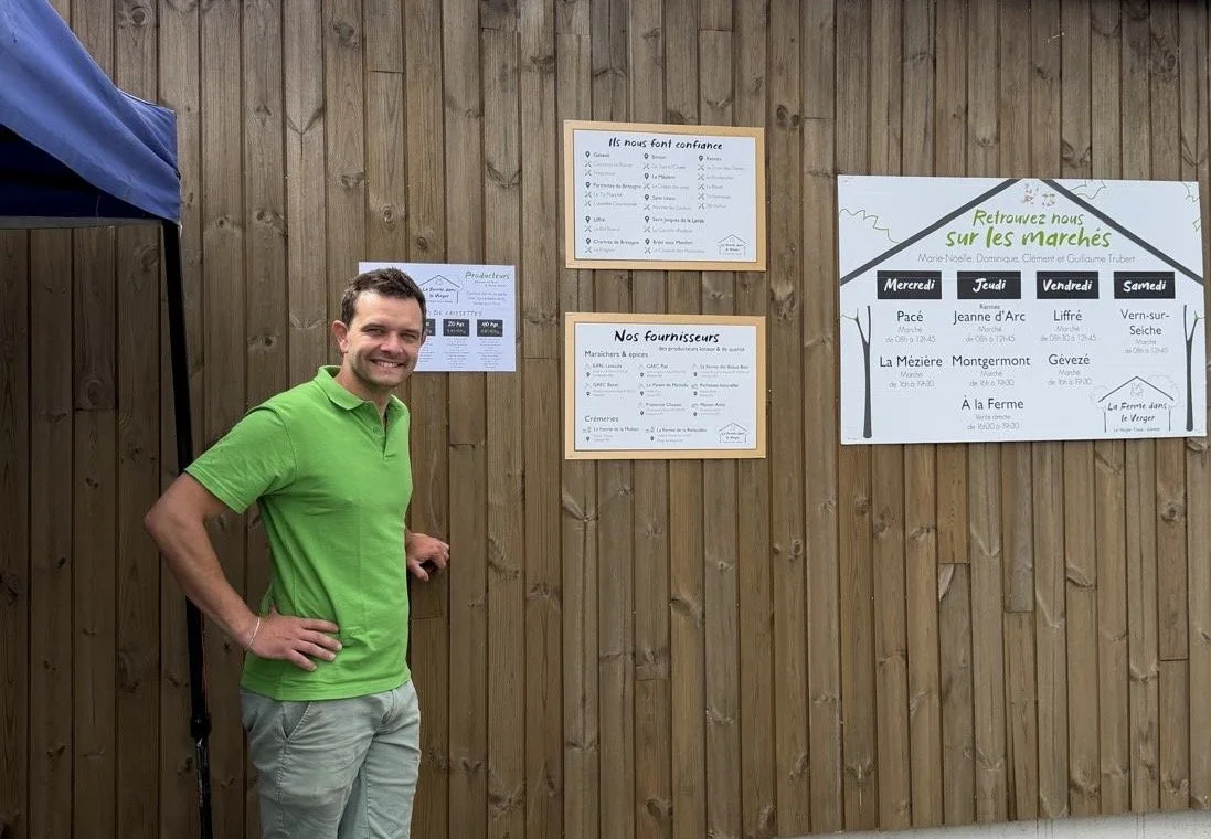 Un homme souriant, habillé en polo vert et pantalons beige, se tient devant un panneau d'affiches en bois avec des informations écrites en français, dans un cadre extérieur.