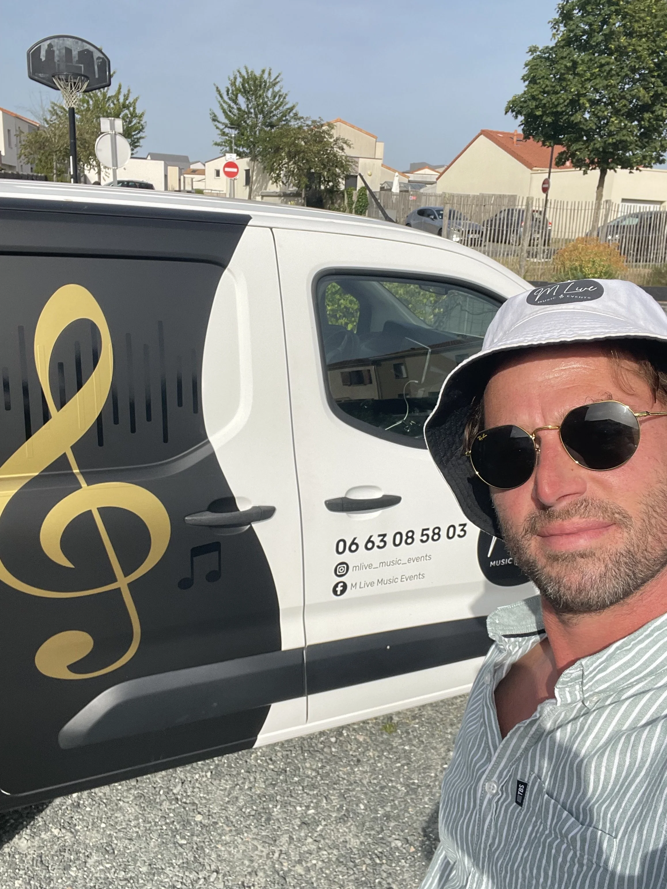 Un homme portant des lunettes de soleil, un chapeau blanc et une chemise à rayures, prenant une selfie devant un véhicule de service pour événements musicaux. Le véhicule affiche un logo avec une clé de sol, ainsi que des informations de contact et des réseaux sociaux.