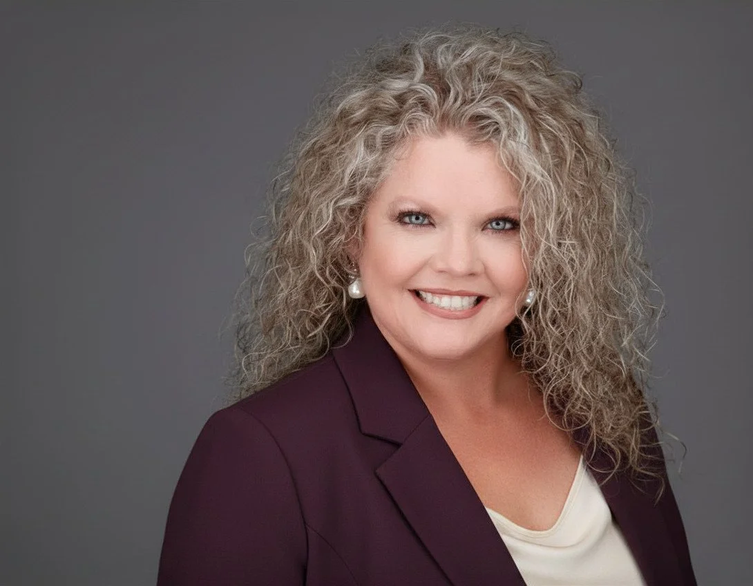 A woman with curly blonde hair, wearing pearl earrings, a maroon blazer, and a white top, smiling against a gray background.
