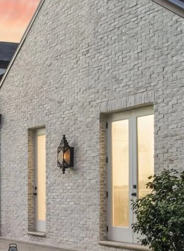 White brick exterior wall with two doors, one smaller and one larger, both with glass panels, and a black exterior wall-mounted lantern.
