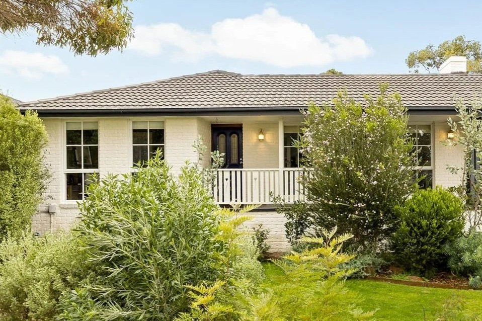 Front view of a limewashed brick house with a tile roof, surrounded by lush green bushes and trees.