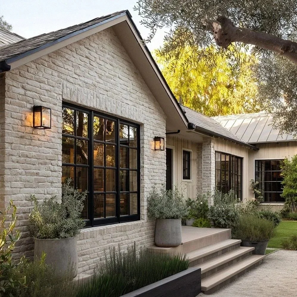 Exterior view of a house with limewashed brick and wood siding, large window with black framing, and outdoor step leading to the entrance, surrounded by potted plants and greenery, with trees and sunlight in the background.