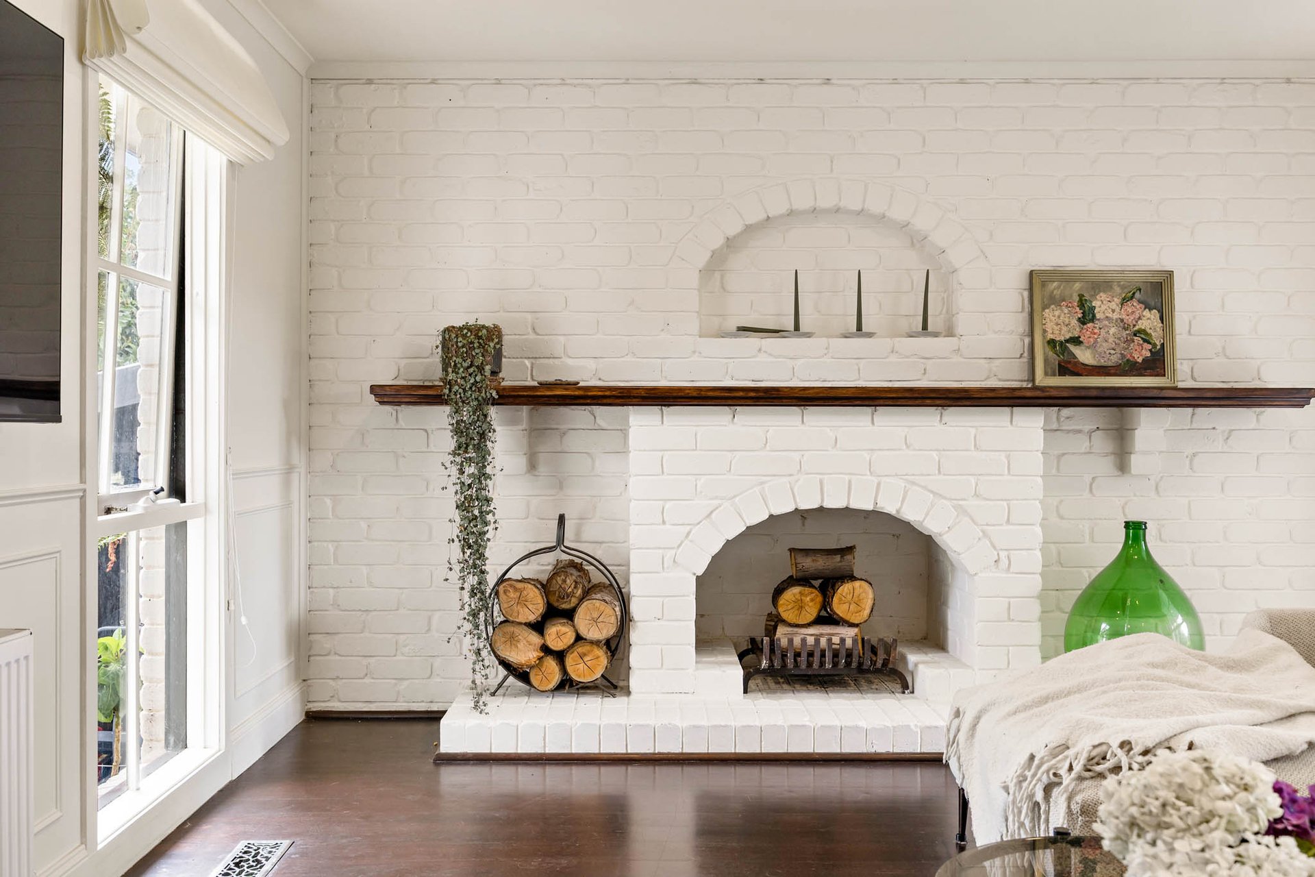 Living room with limewash brick fireplace, wood logs, and a framed floral painting on the mantel, next to a green vase and a window with white curtains.