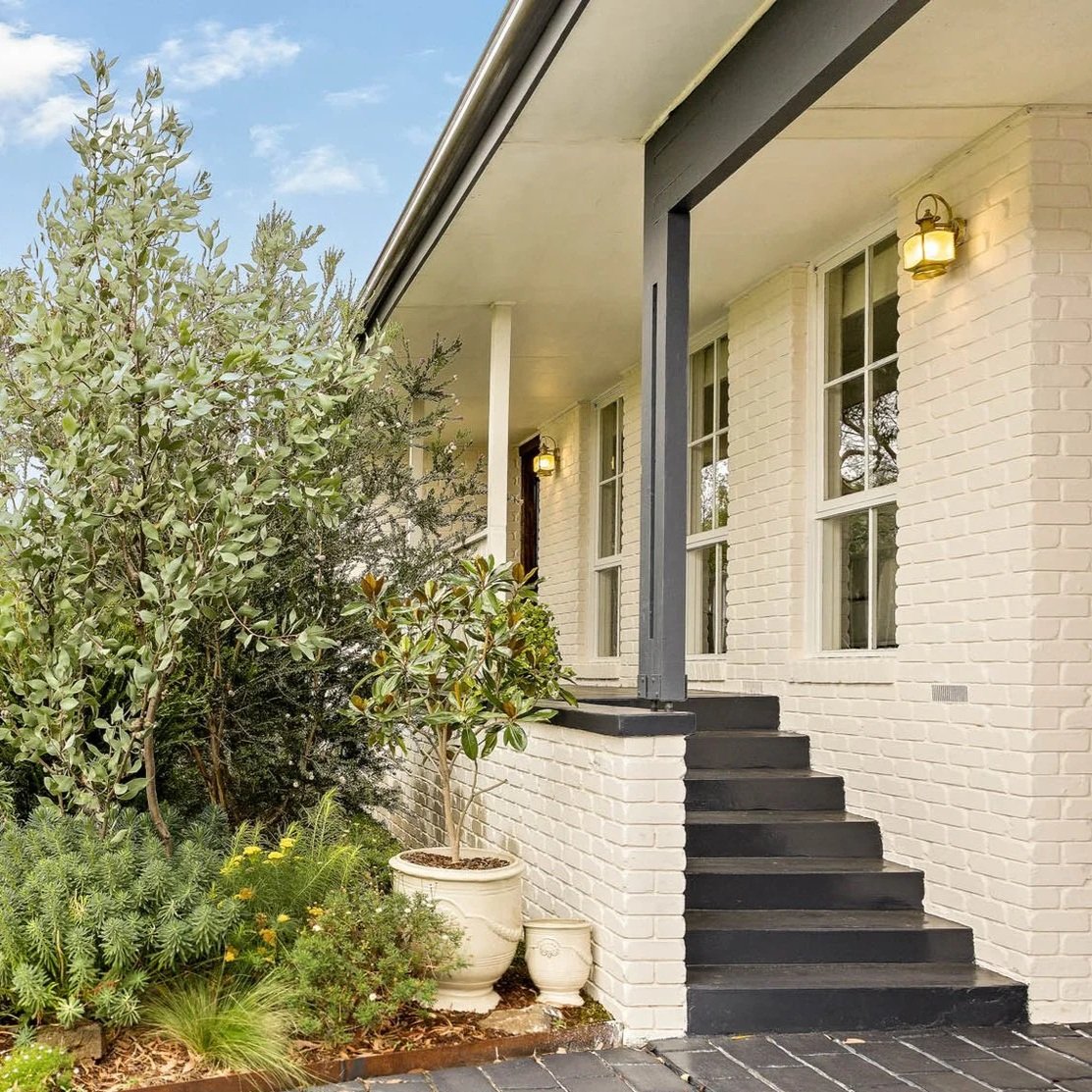 Close-up of the front porch of a white brick house with black stairs, potted plants, and greenery in the garden.