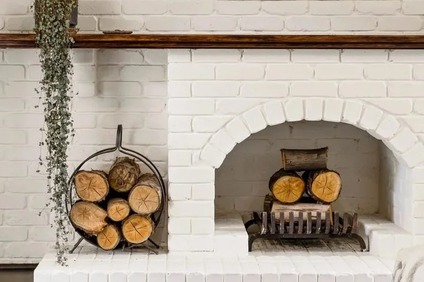 Wood logs stacked in a firewood holder and inside a brick fireplace with a decorative black metal grate.