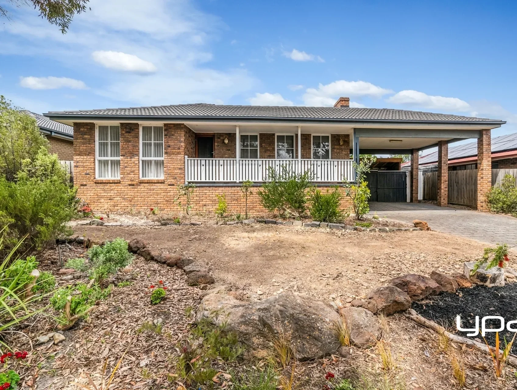 A brick house with a front porch and a driveway, surrounded by sparse landscaping with some plants and rocks, under a blue sky with scattered clouds.