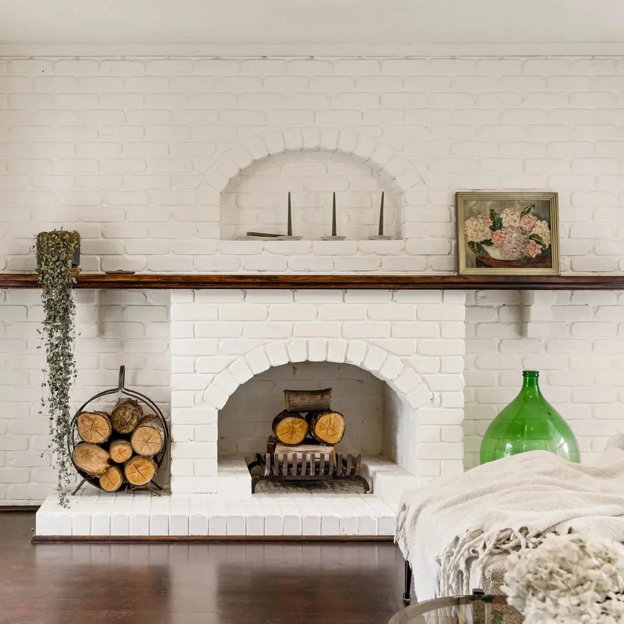 Living room with limewashed brick fireplace, logs stored in a metal holder, a wooden mantel with candles and a floral painting, green glass bottle, and a beige throw blanket on furniture.