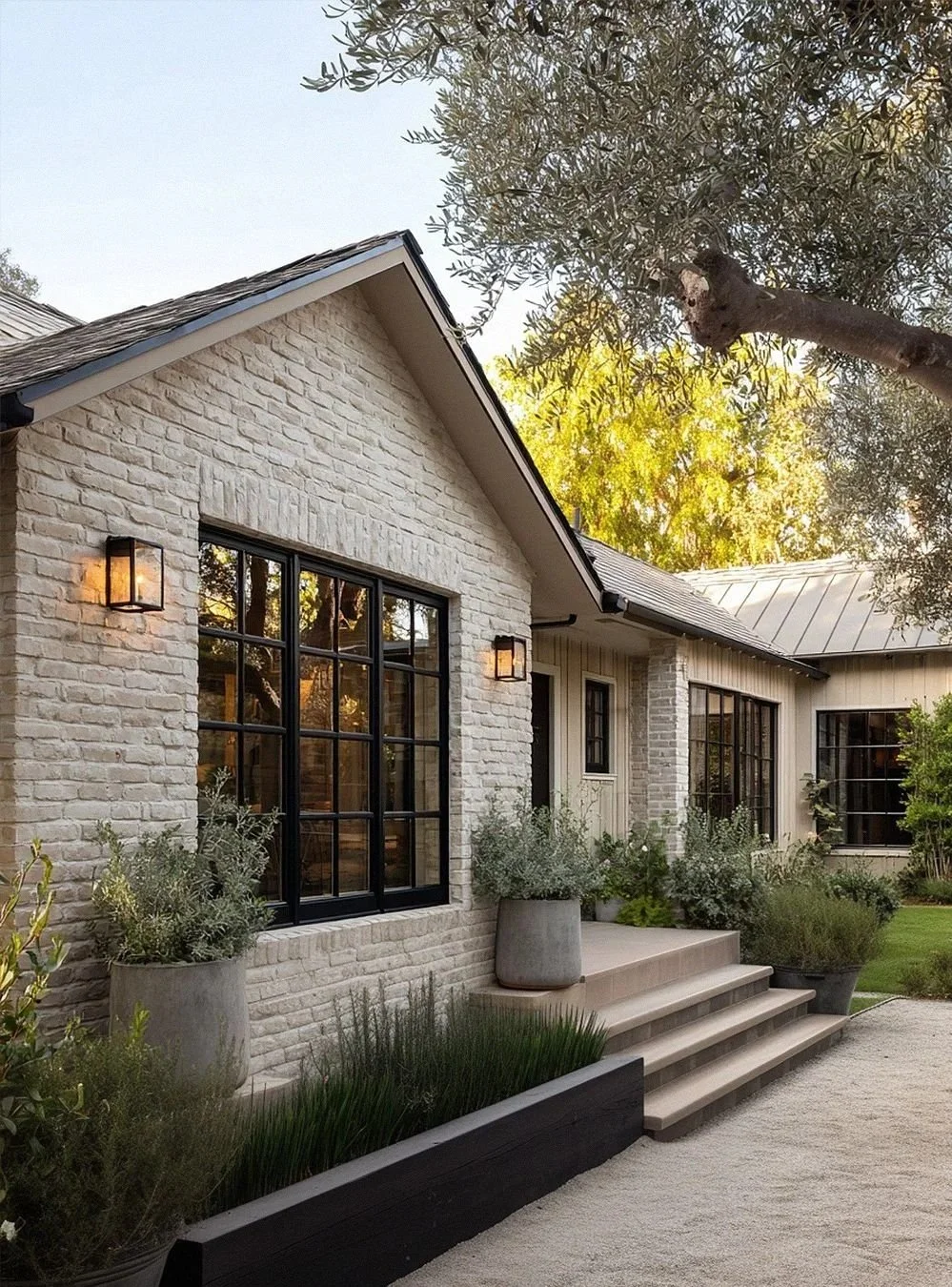 Exterior of a modern house with limewashed brick walls, black-framed windows, and a small set of steps leading to the entrance. There are potted plants and greenery near the steps, and outdoor wall-mounted lights are on either side of the window.