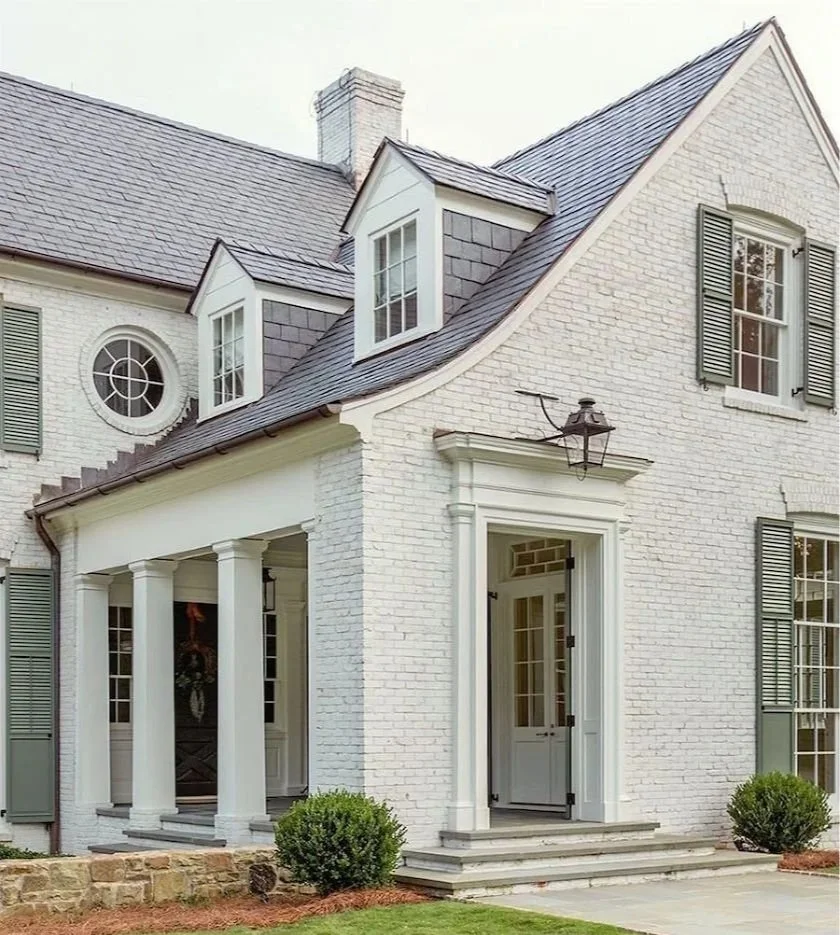 A limewashed brick house with a gabled roof, dormer windows, and an entrance with columns and steps, surrounded by shrubs.