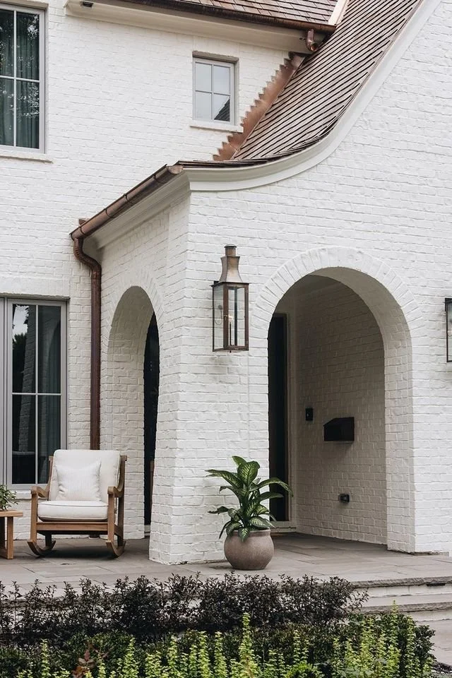 Front porch of a whitewashed brick house with arched entryway, a hanging lantern, potted plant, outdoor chair, and garden plants.