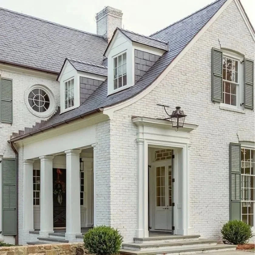 Front of a limewashed brick house with a porch supported by white columns, storm door, multiple windows with shutters, and dormer windows on the roof.