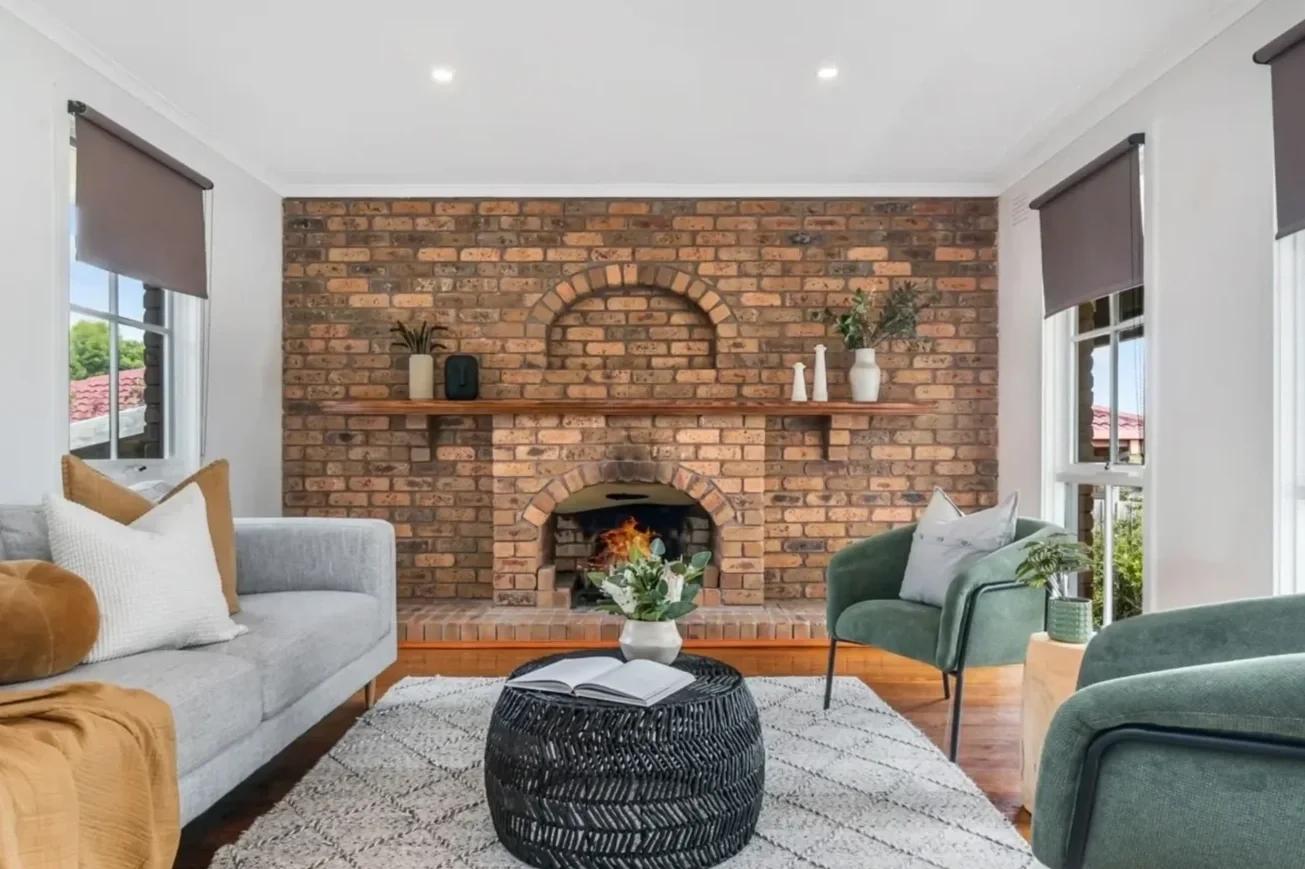 Living room with a brick fireplace, a gray sofa, green armchairs, a black woven coffee table with an open book and a flower arrangement, and windows with gray blinds.