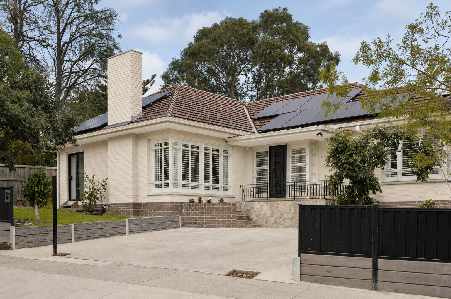 A 60s limewashed brick house with solar panels on the red tile roof, a chimney, large windows with white shutters, a front porch with a black gate, and a driveway with a black fence in a suburban neighborhood.