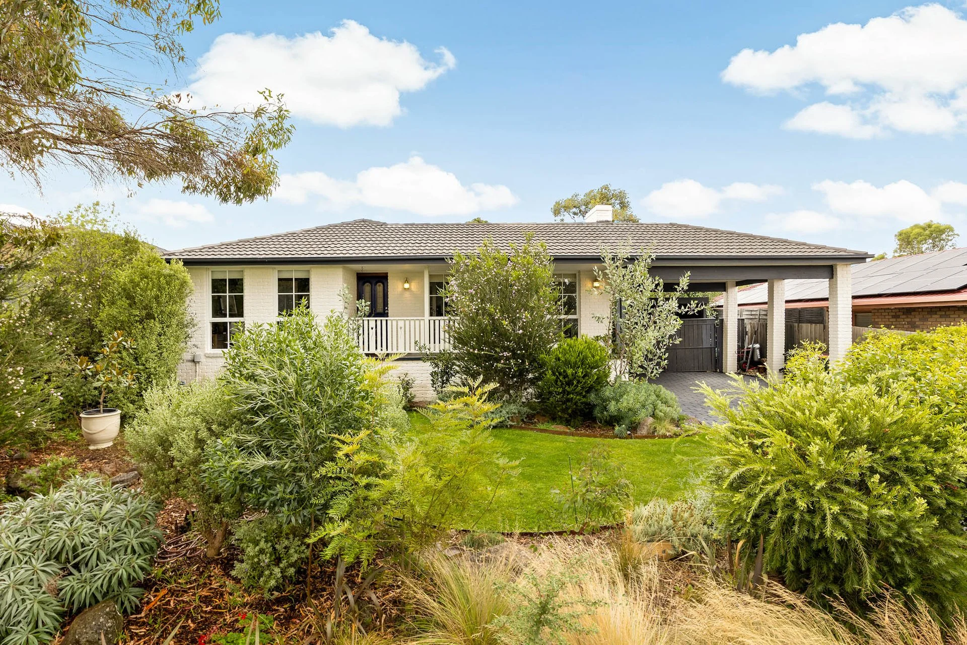 A single-story Limewashed house with a front porch, surrounded by lush green garden plants and trees, under a bright blue sky with scattered clouds.