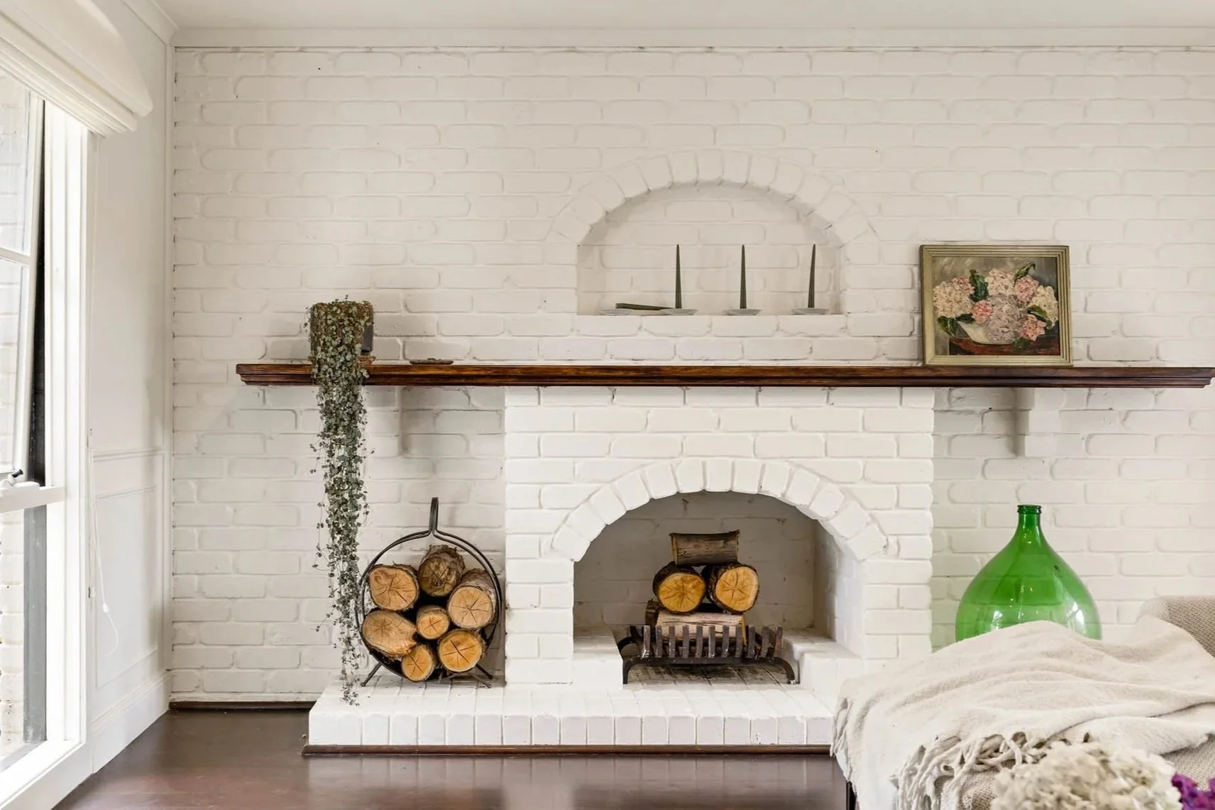 White brick fireplace with logs inside, wooden mantel with decorative items including candles and a framed floral painting, and a green glass vase to the right. A armrest with a blanket or throw is partially visible in the foreground.