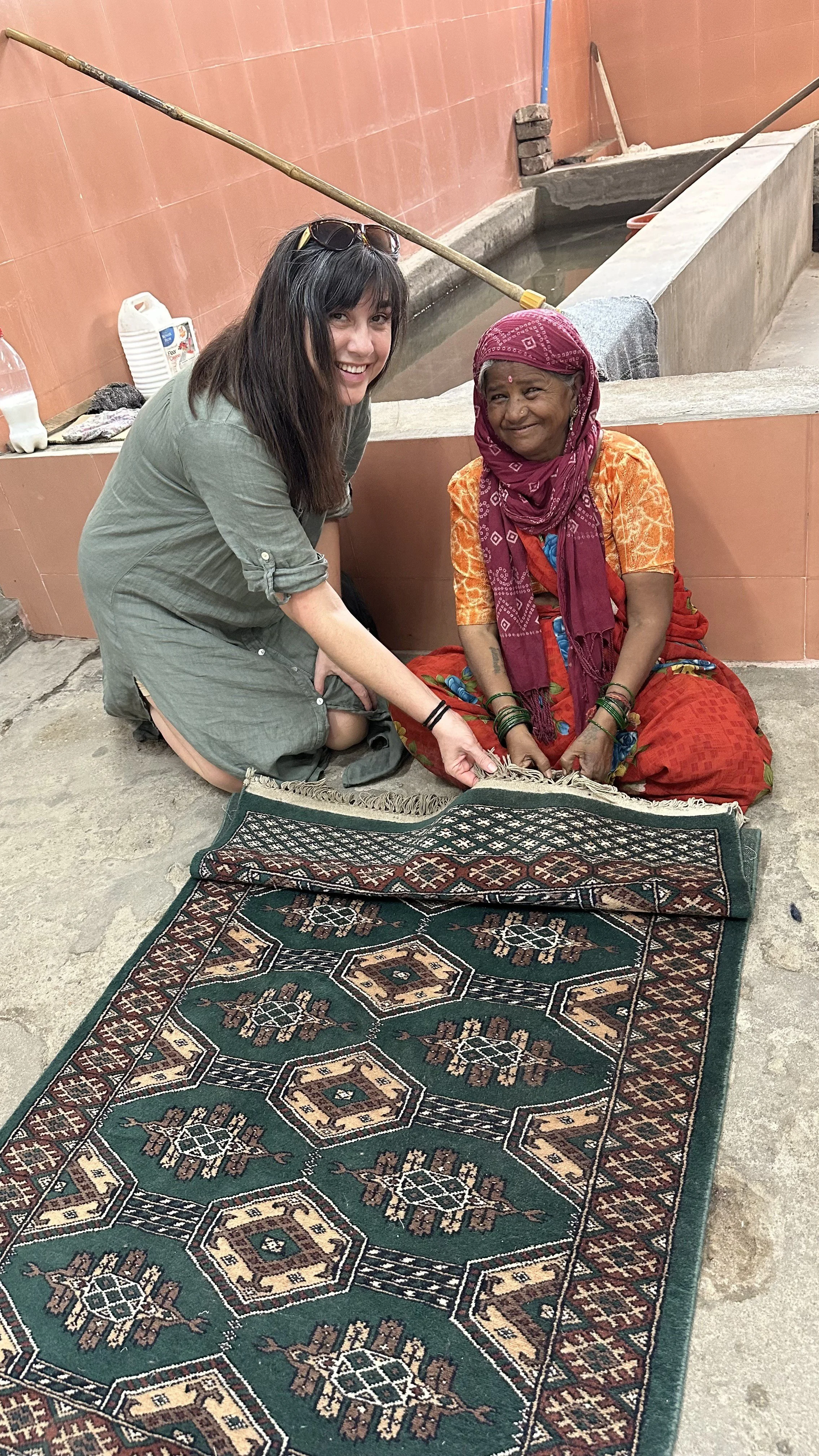 A woman is sitting next to an older woman on the floor, holding a patterned rug. They are near a tiled wall and a water tank, with various objects around, including bottles and tools.
