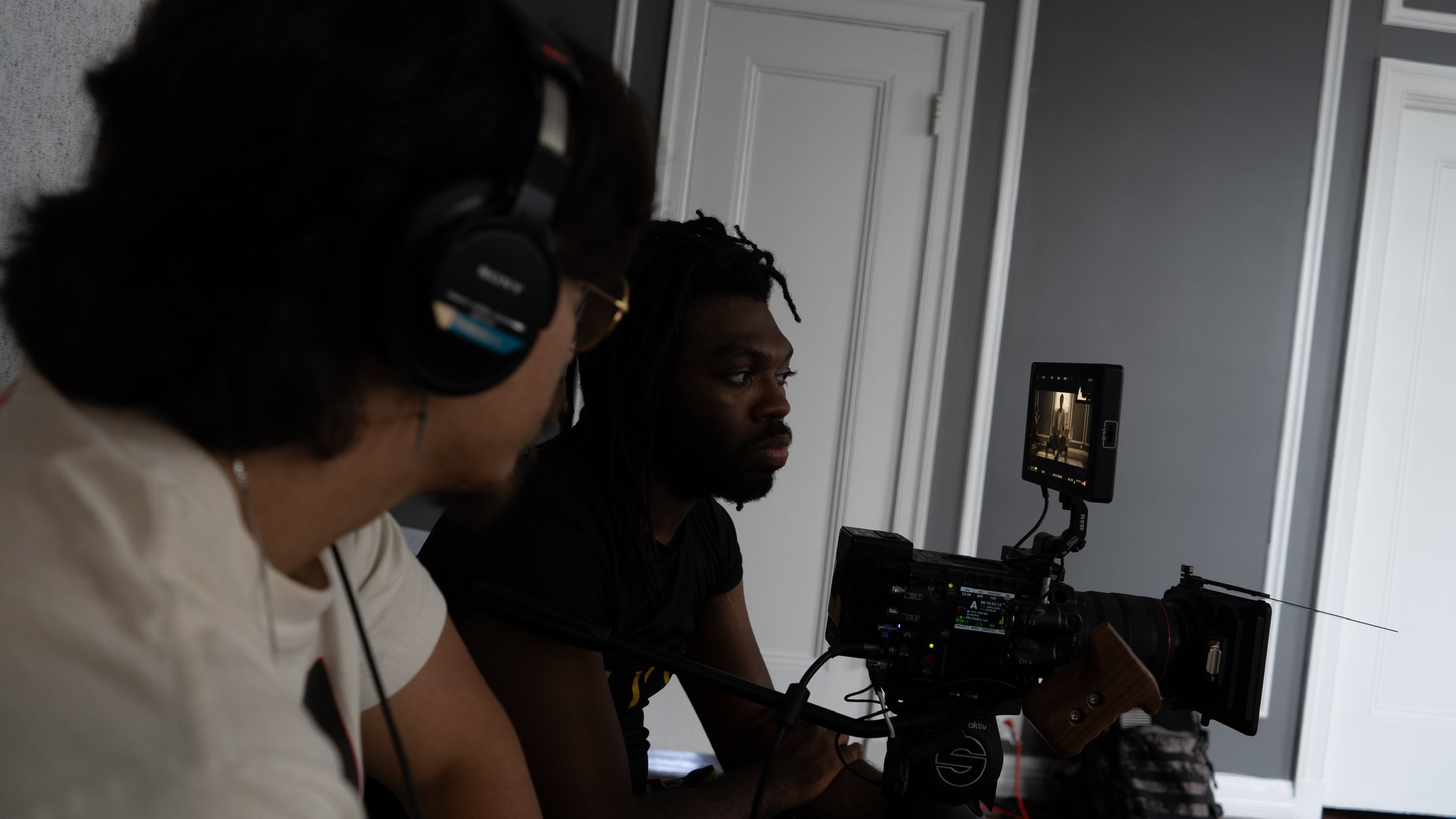 Two people working on a film or video shoot indoors, one person operating a professional camera and the other wearing headphones and looking at a monitor.