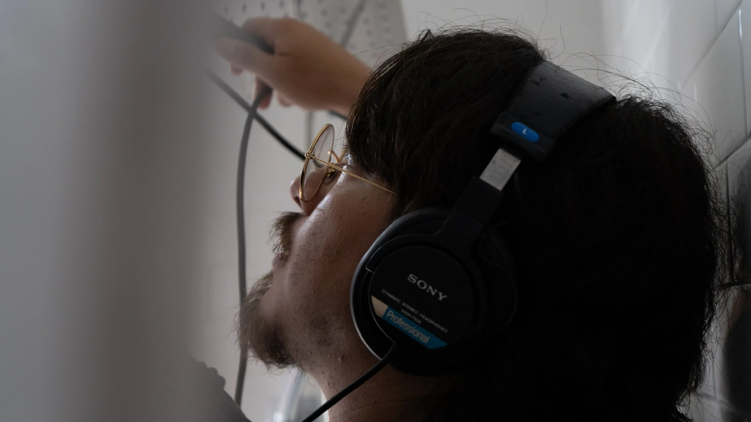 Side view of a man wearing glasses and Sony headphones, resting with his head against a wall, holding camera equipment above his head.
