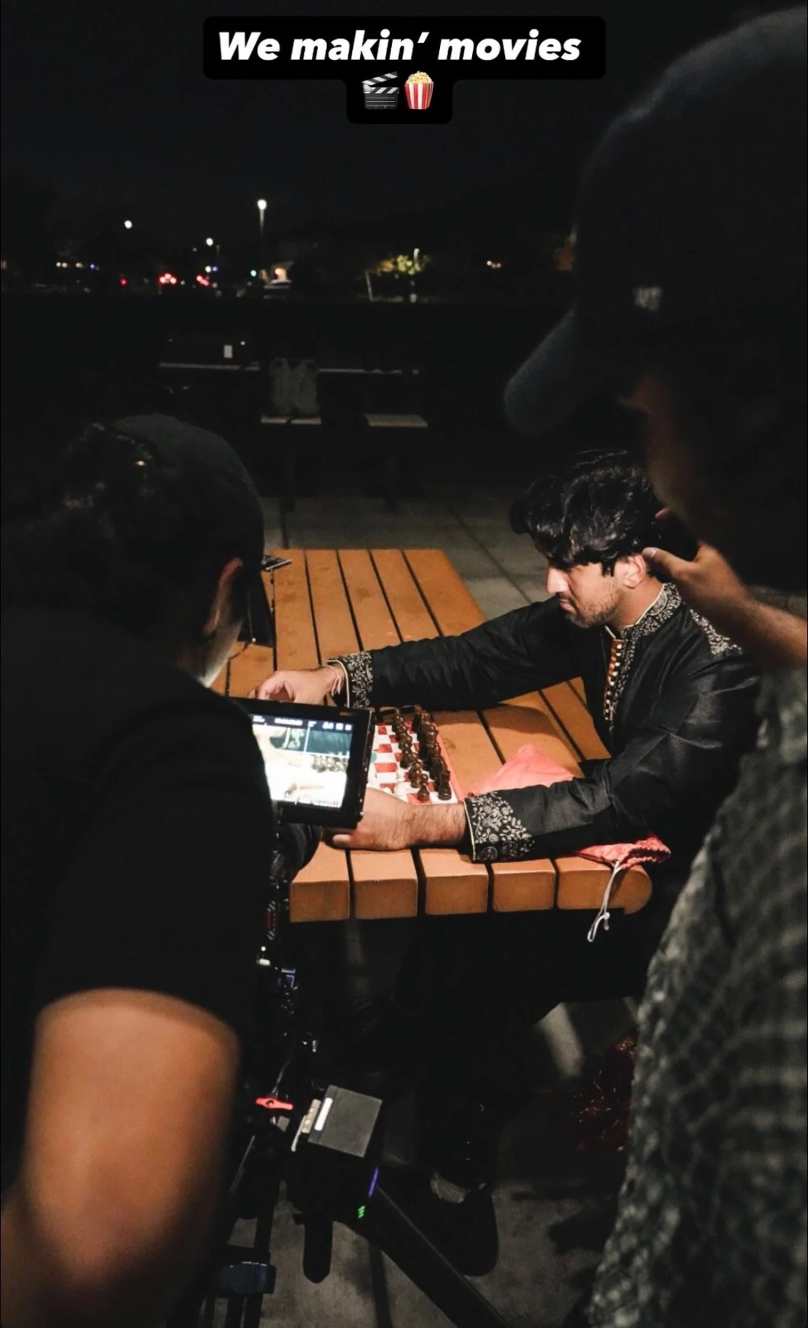 People filming a man playing chess at night at an outdoor table.