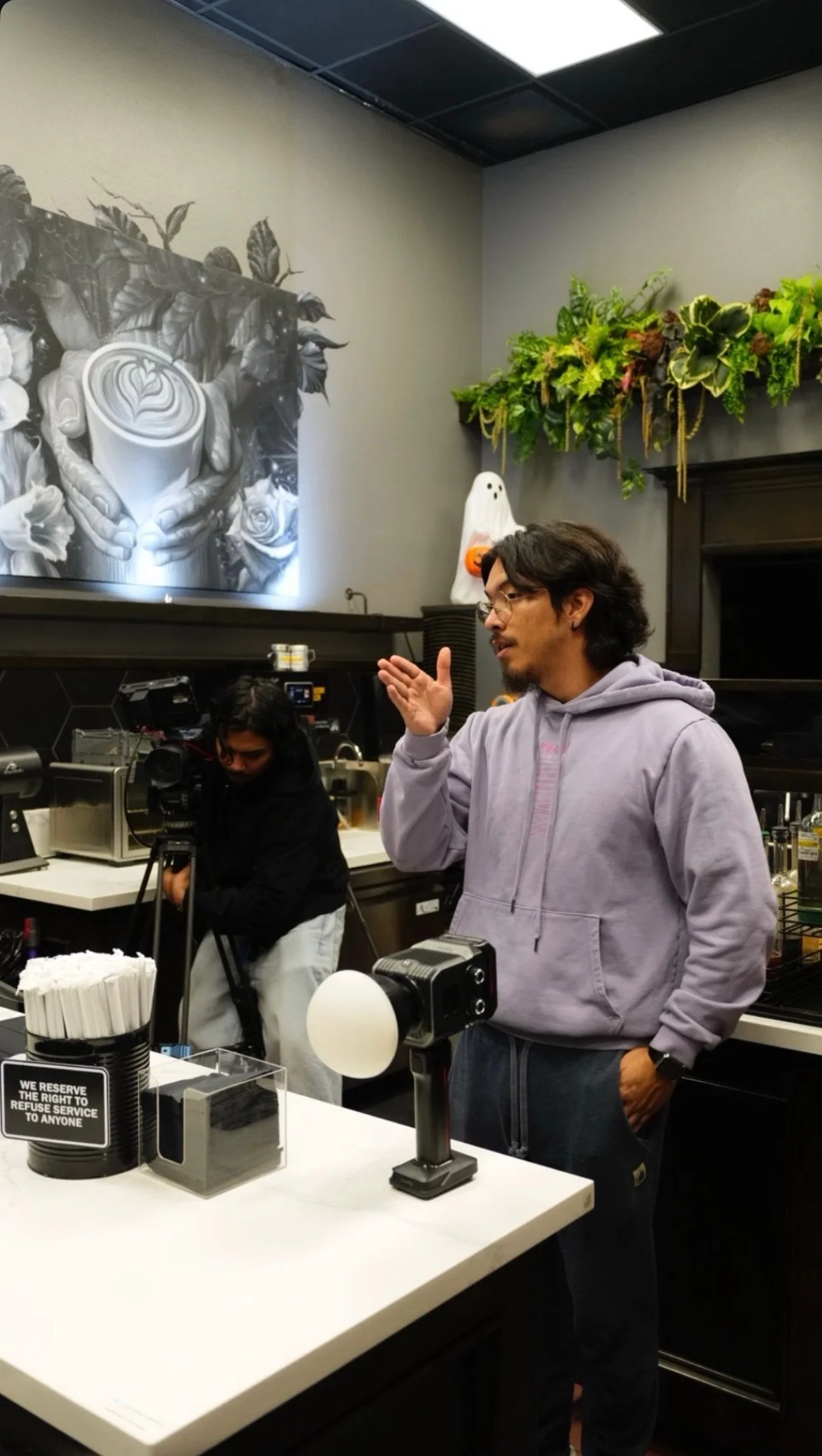 A man with glasses and a beard wearing a lavender hoodie is speaking or explaining something in a restaurant or cafe. Behind him, a woman with dark hair is sitting and appears to be working with a camera. There is a Halloween-themed ghost decoration 