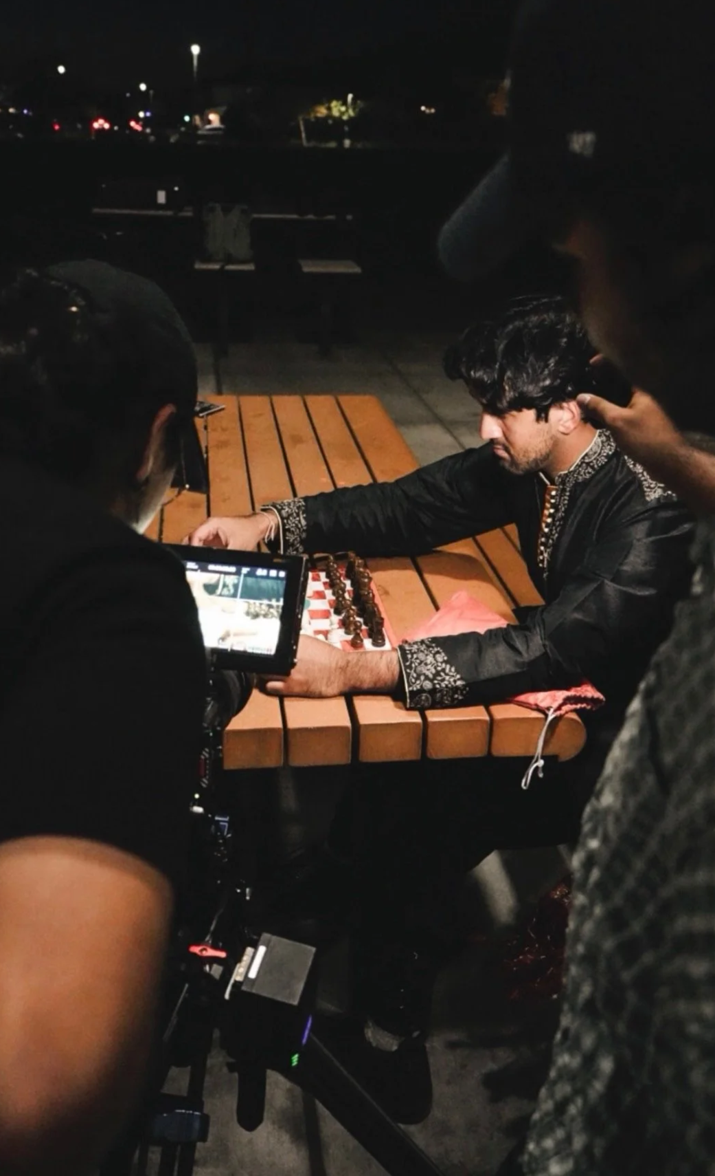 A man in traditional black attire with embroidered details plays chess outdoors at night, surrounded by people filming him, near a wooden table with a pink cloth and a chessboard, with city lights and streetlights in the background.