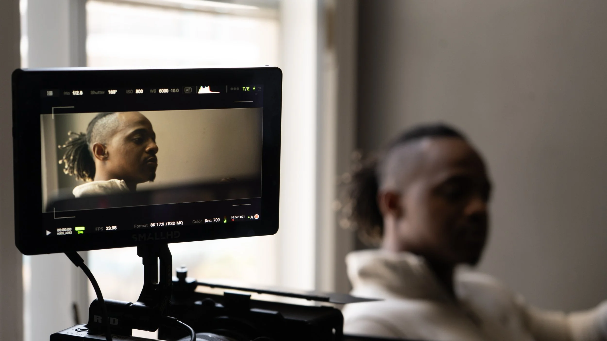 Video monitor showing a woman with dreadlocks appearing in camera view, with a person blurred in the foreground, in a room with natural light.