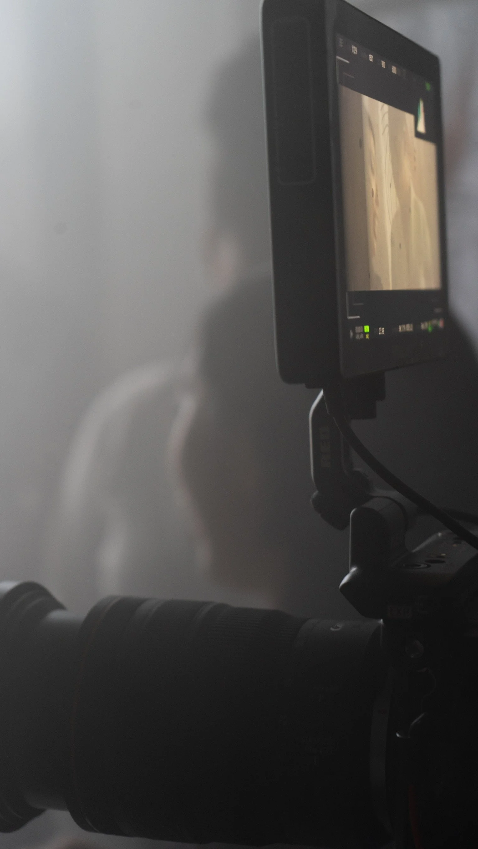 Close-up of professional camera equipment, including a camera monitor and a zoom lens, with a plain gray background.