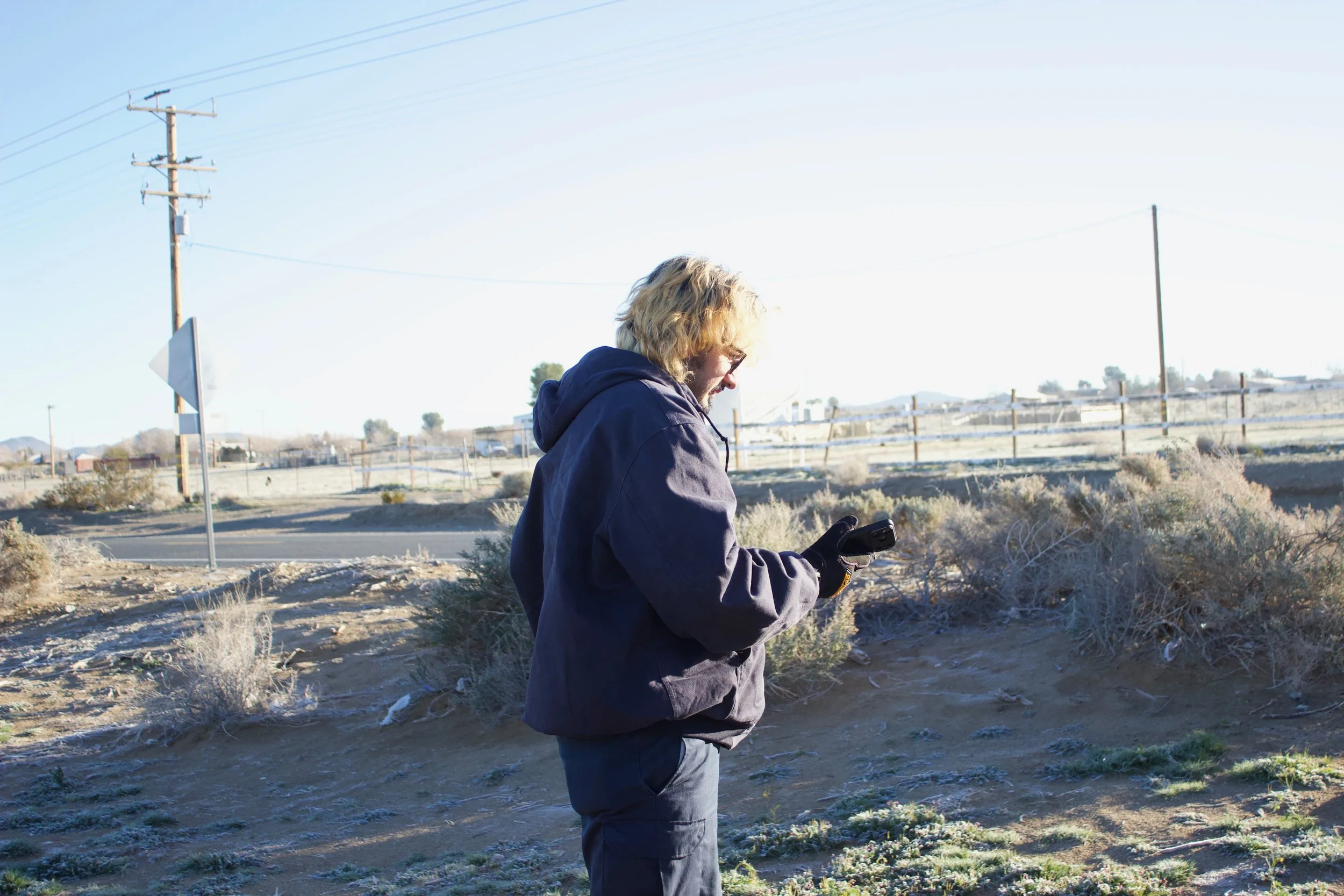 A man with Blonde hair, glasses, wearing a dark hoodie and gloves who is outdoors near a desert area with power lines in the background.