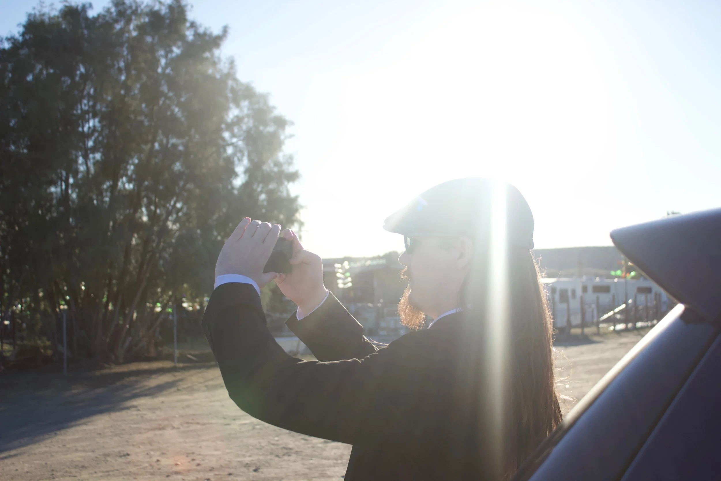 Person in formal attire taking a photo with a smartphone outdoors during daytime, with sunlight and trees in the background.