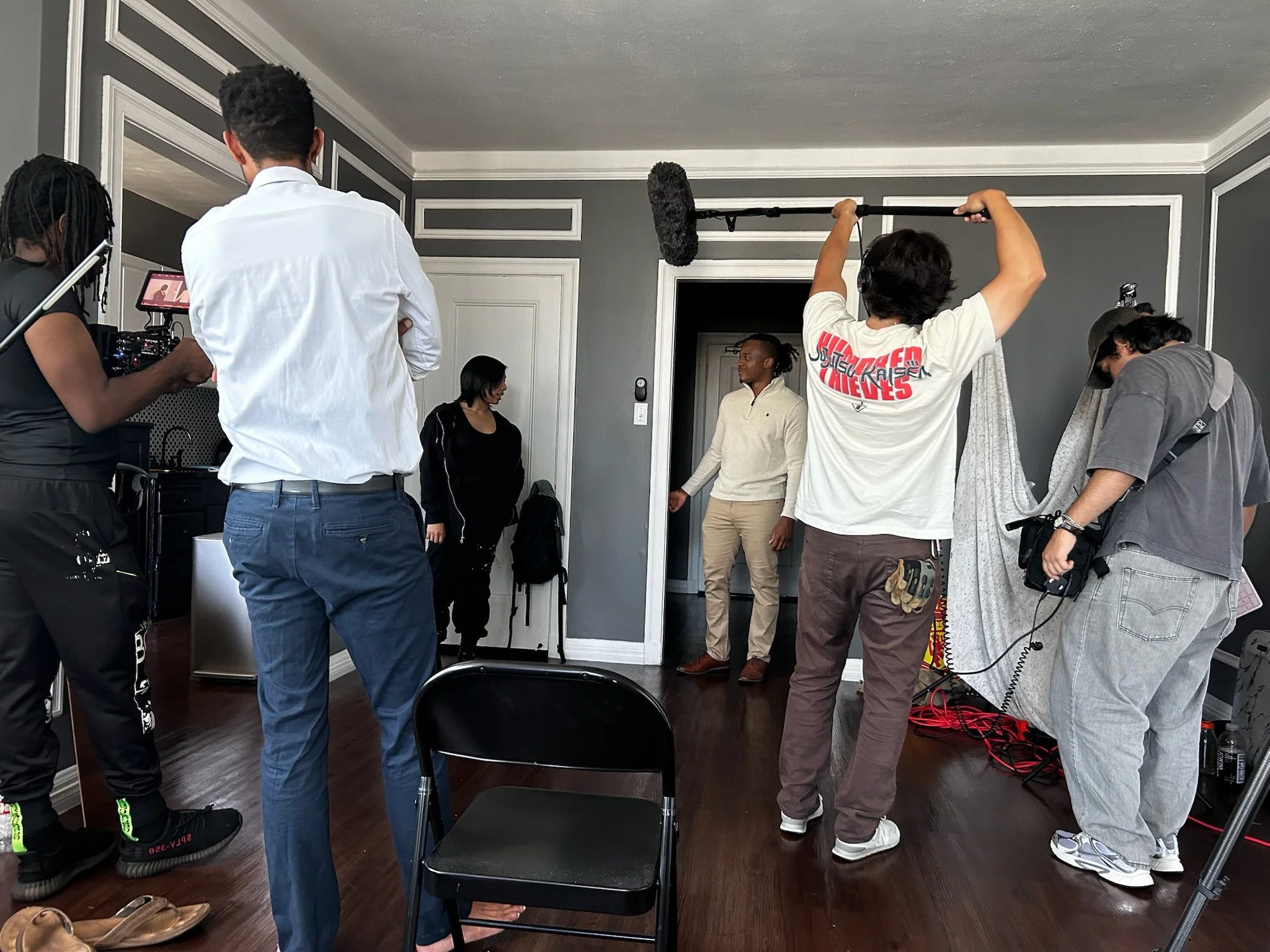Behind-the-scenes scene from a video or film shoot with crew members and equipment, including a boom mic, camera, and lighting, in a room with dark hardwood floors and gray walls with white trim.
