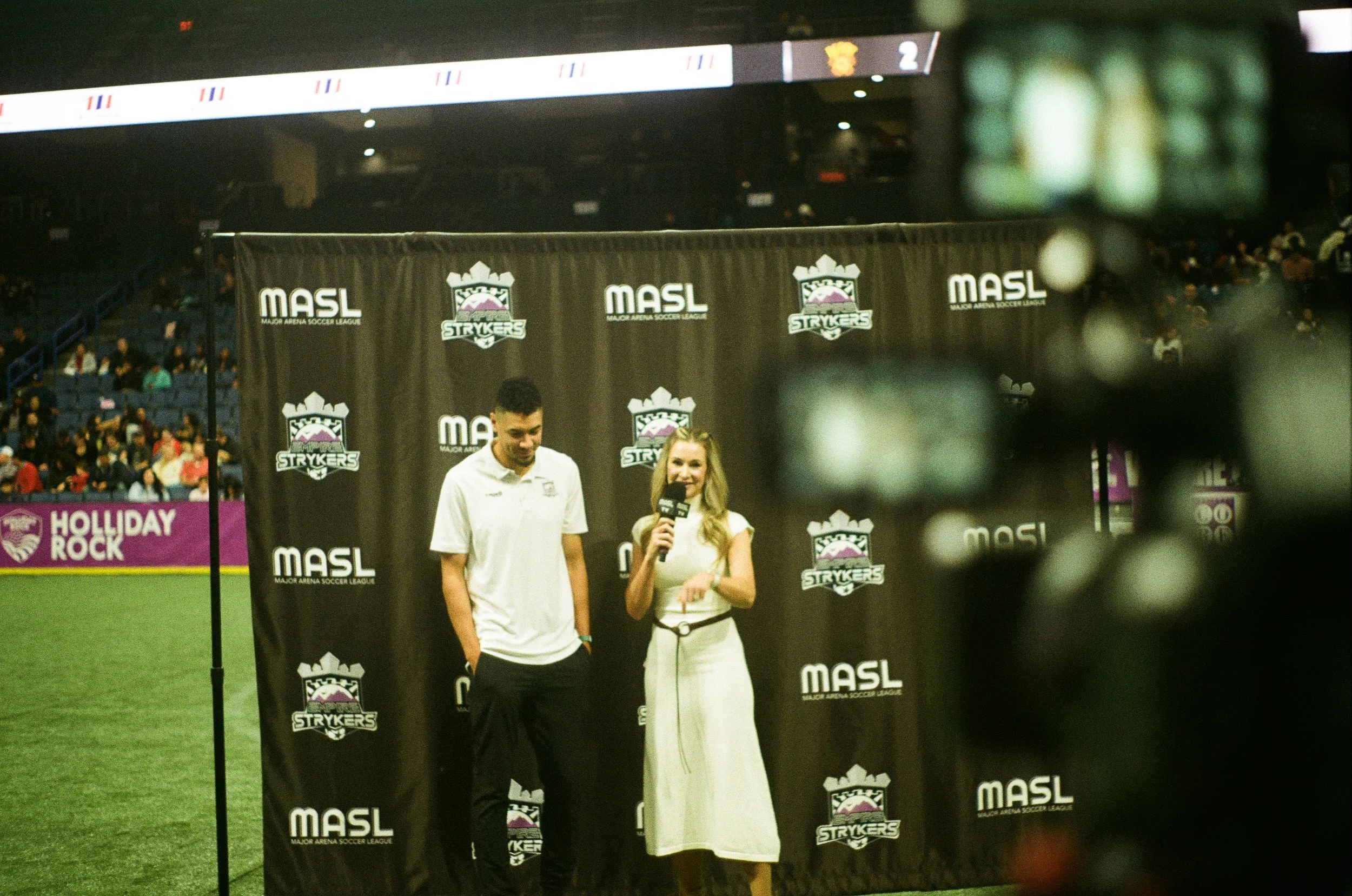 Two people standing behind a black backdrop with the logos of MASL Major Arena Soccer League and Strykers, one man and one woman, on a soccer field with an audience in the background.