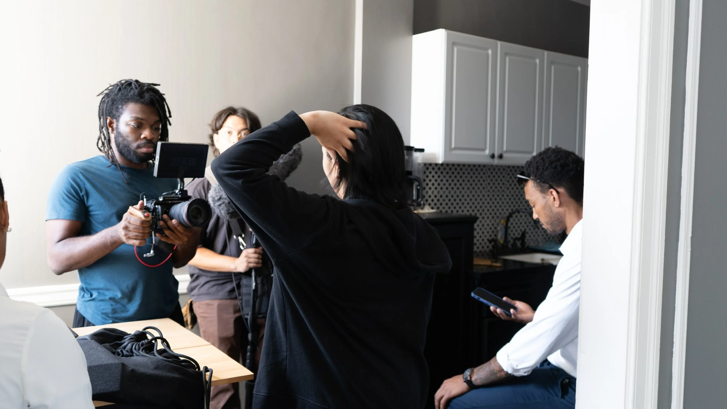 A group of people on a film or video production set, with one person holding a camera, a woman adjusting her hair, and others engaged in discussion or using their phones in a kitchen setting.