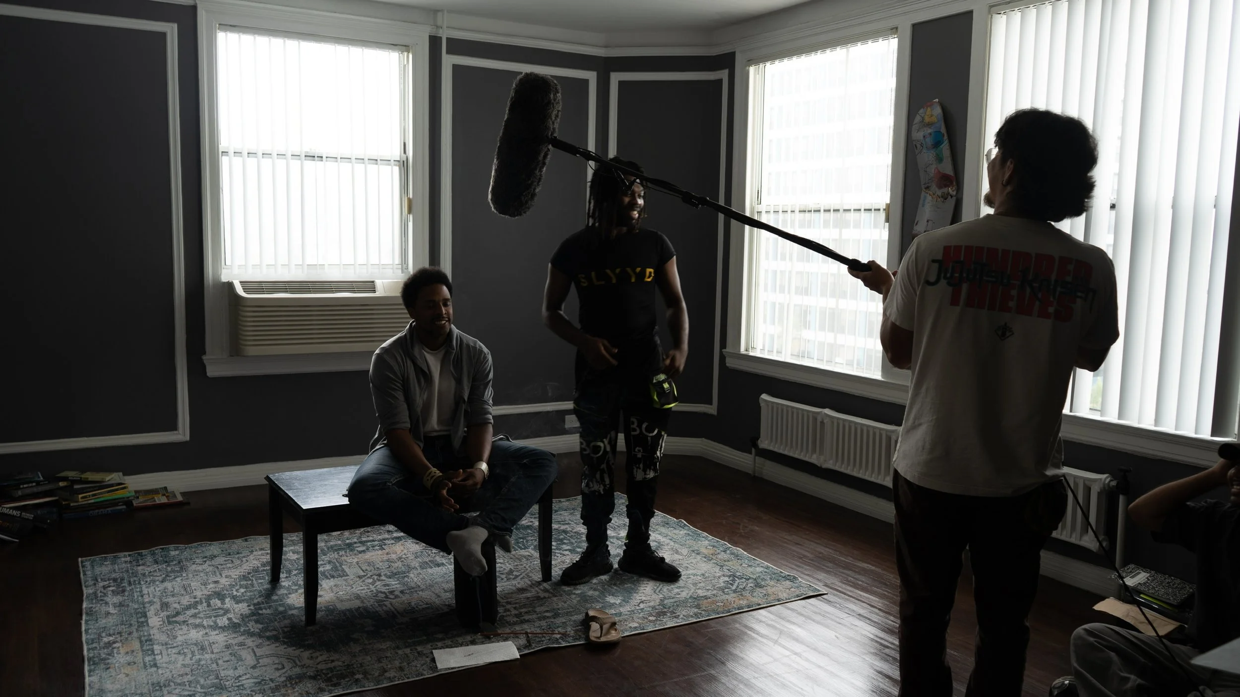 A video or interview being filmed inside a room with three people. One person is sitting on a chair, another person is standing and smiling, and a third person is holding a boom microphone over their head. The room has large windows with blinds, a bo
