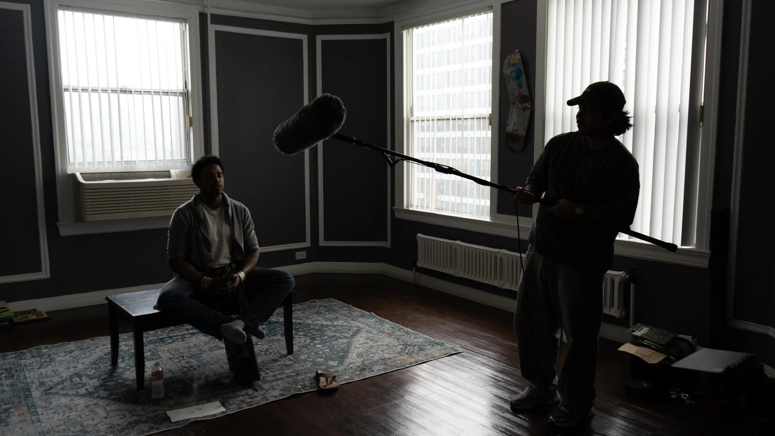 Interior of a room with dark walls and large windows with vertical blinds, a man sitting cross-legged on a coffee table, and another person operating a boom microphone. There is a skateboard hanging on the wall and a carpet on the wooden floor.