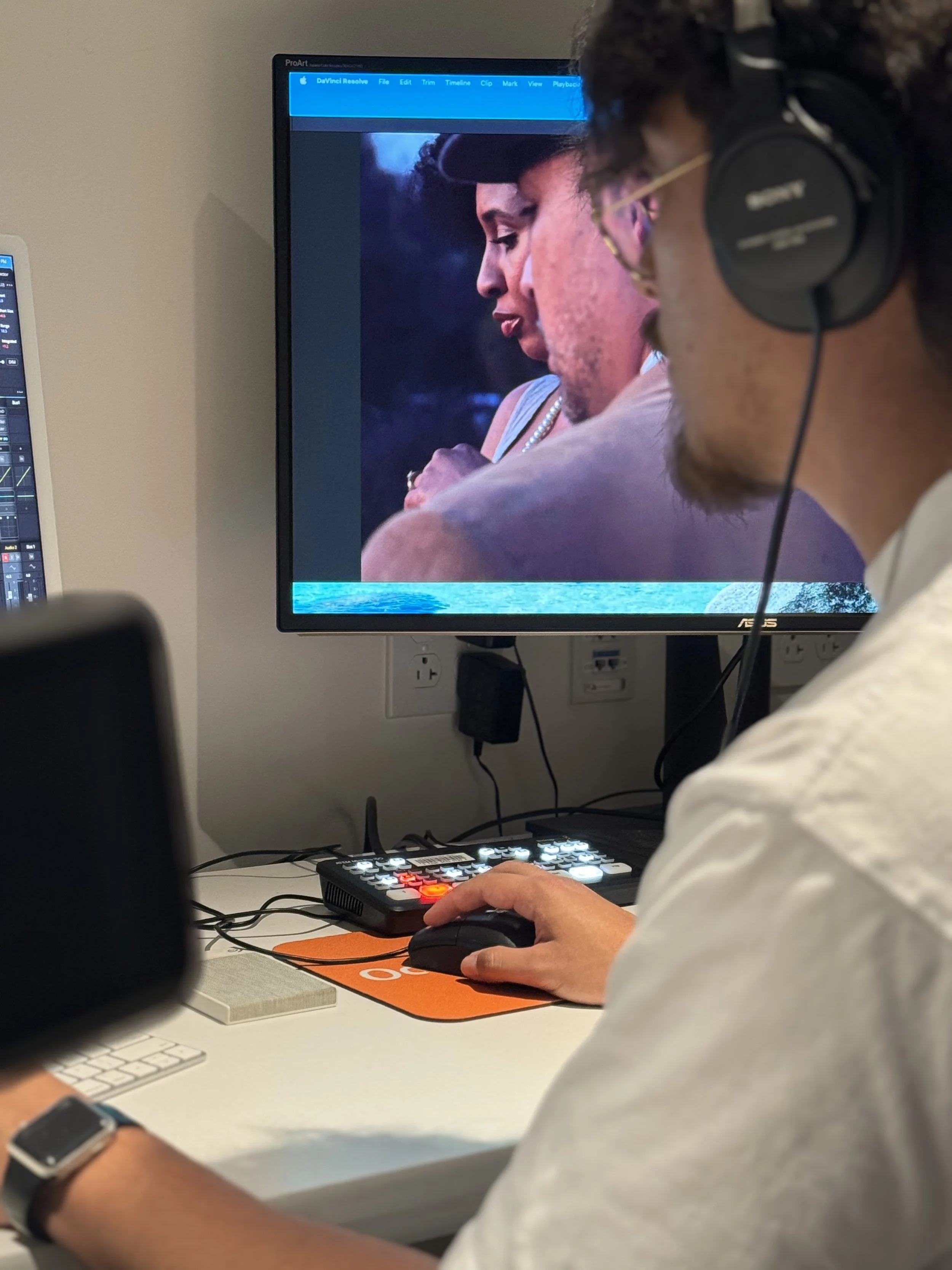 The monitor displays an image of two individuals in profile, one wearing a hat and jewelry, with a nature background. The person's hand is on a mouse and they are sitting at a white desk with a keyboard, a computer mouse, and a desk mat.
