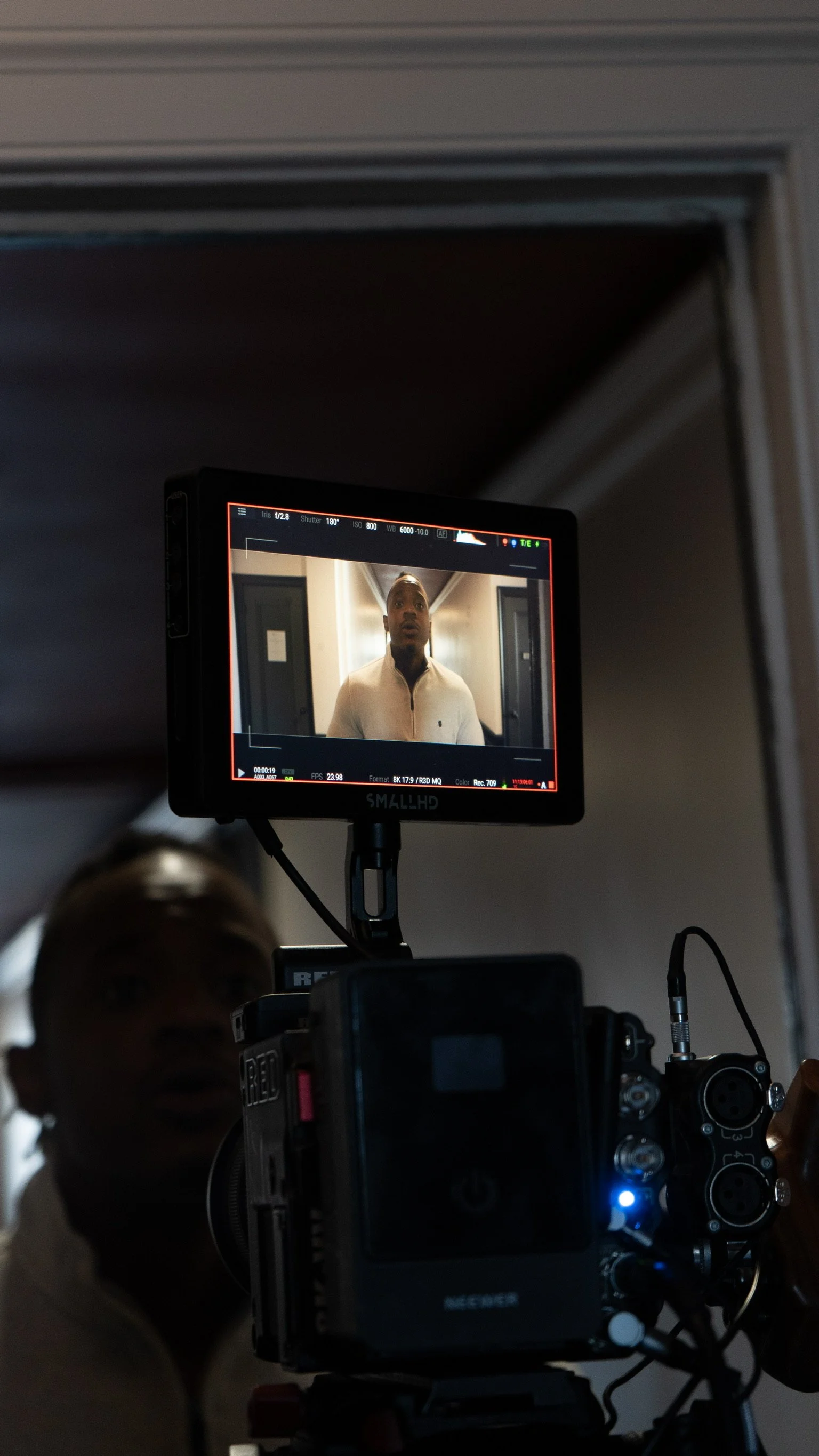 On a film set, a camera and monitor display a man in a beige sweater standing in a hallway, preparing for filming.