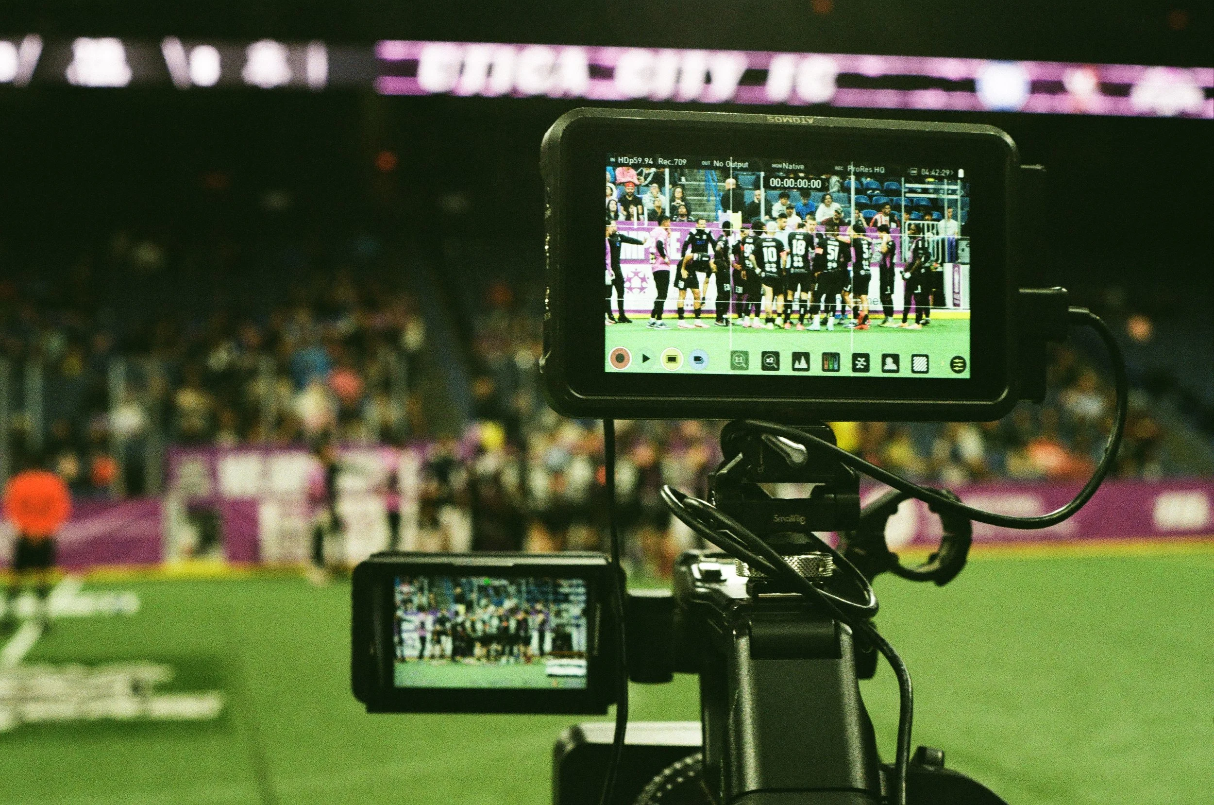 A camera or recording device capturing a sports event on a field in a stadium filled with spectators.