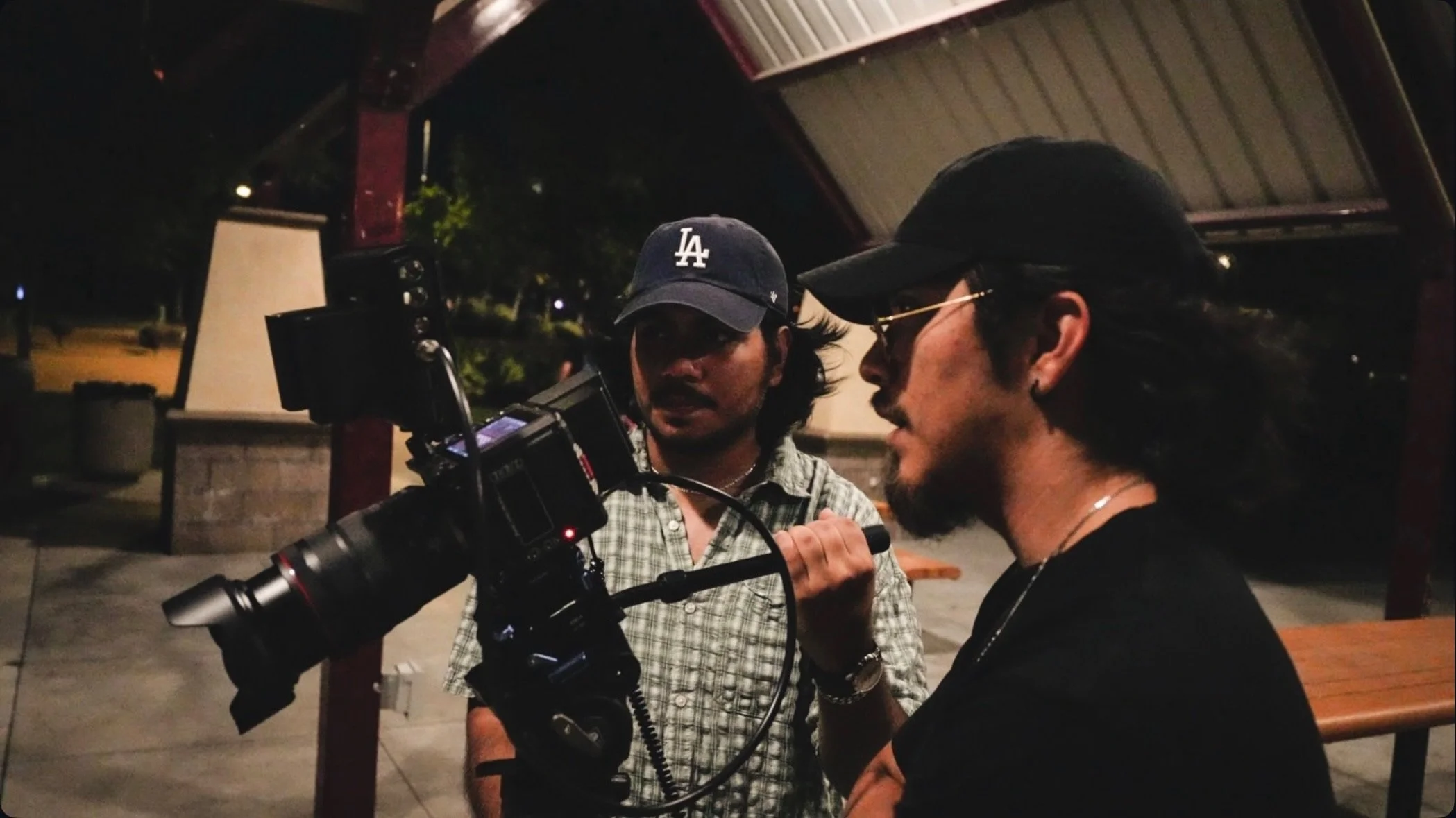 Two men, one with a camera and the other with glasses and a black cap, are engaged in a conversation at night outdoors under a covered structure.