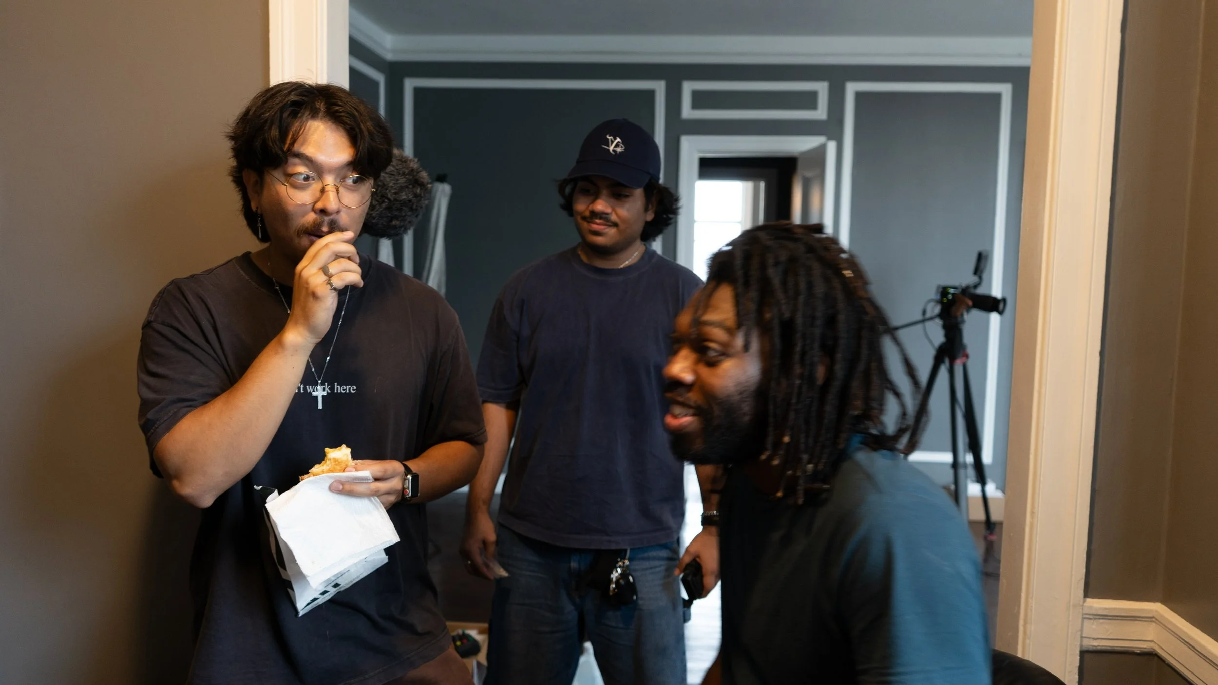 Three men in a room having a conversation, one holding a piece of bread and tissue, with a camera on a tripod in the background.