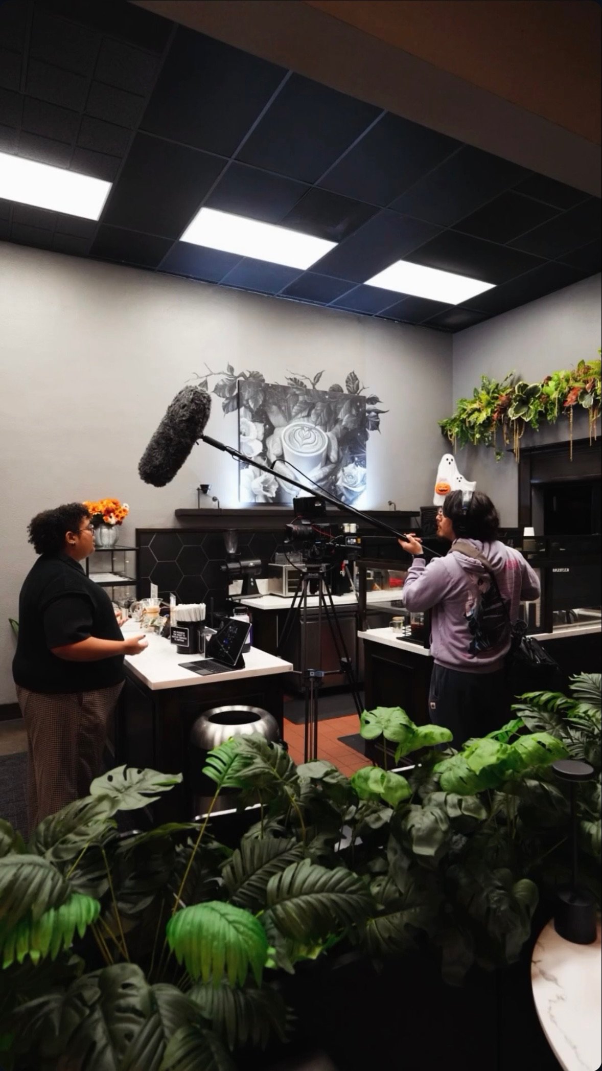 A person being interviewed in a cafe, with a crew member operating a boom microphone, indoor decor with plants and artwork.
