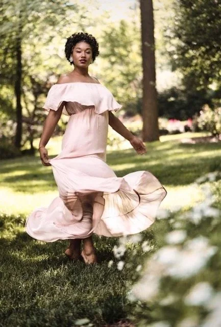 A woman in a light pink off-the-shoulder dress dancing outdoors in a park with trees and grass.
