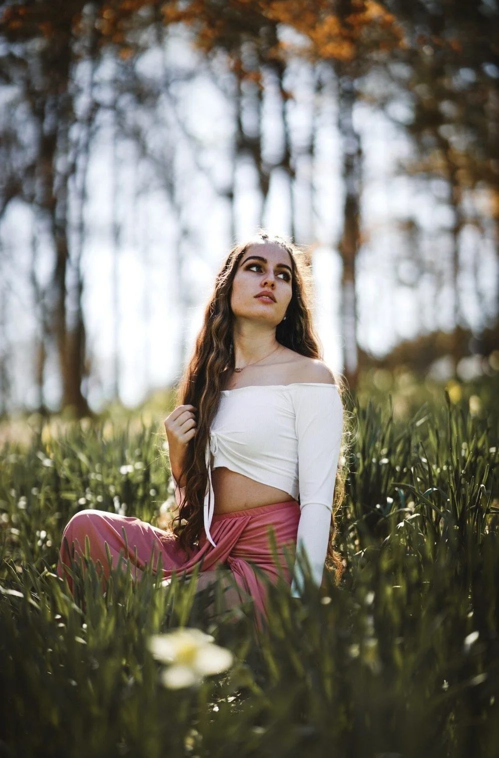 A young woman with long, wavy brown hair sitting in a field of tall green grass during daytime, with tall trees in the background and sunlight filtering through the branches.