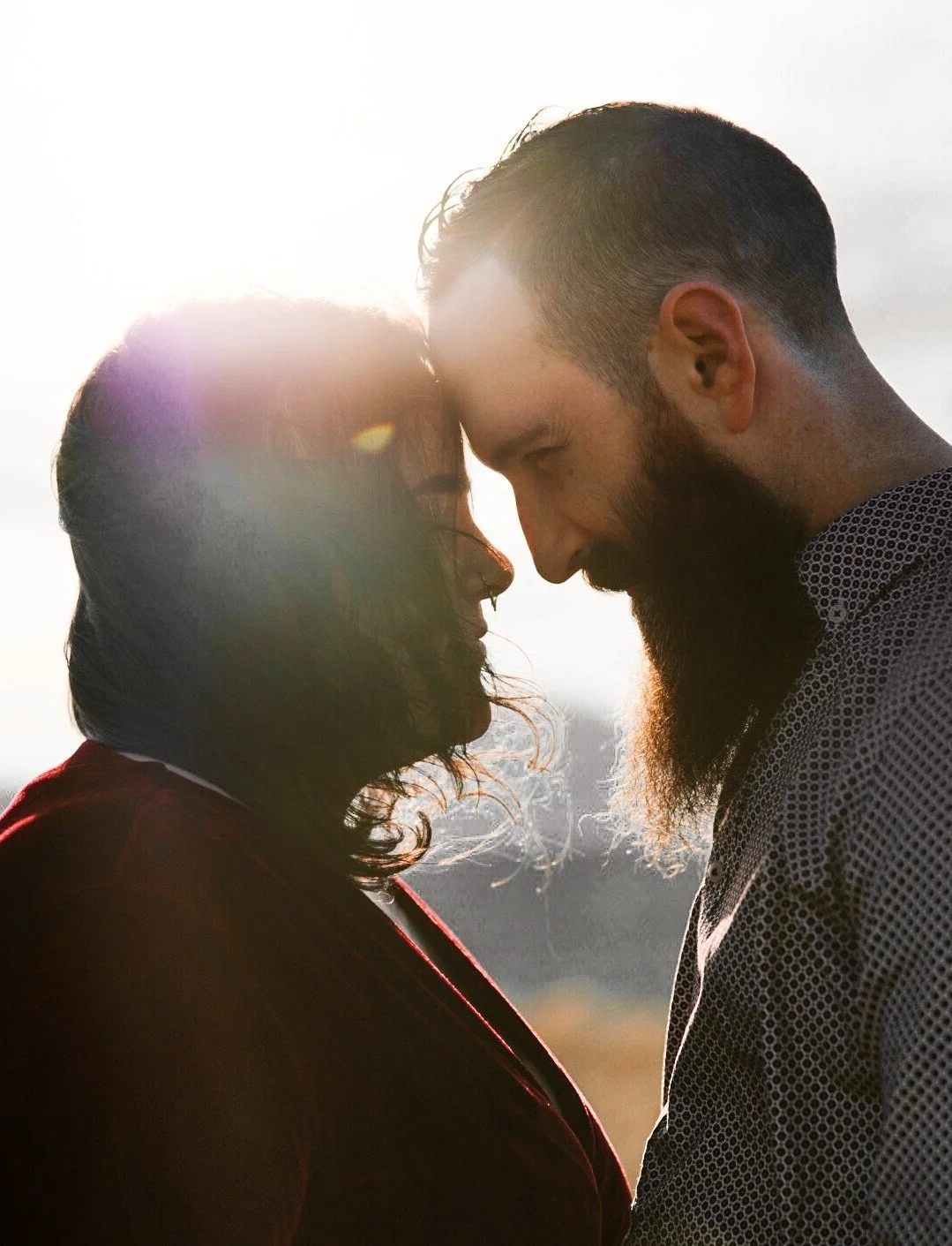 A couple is standing close together with foreheads touching, backlit by the sun, creating a warm glow around them.