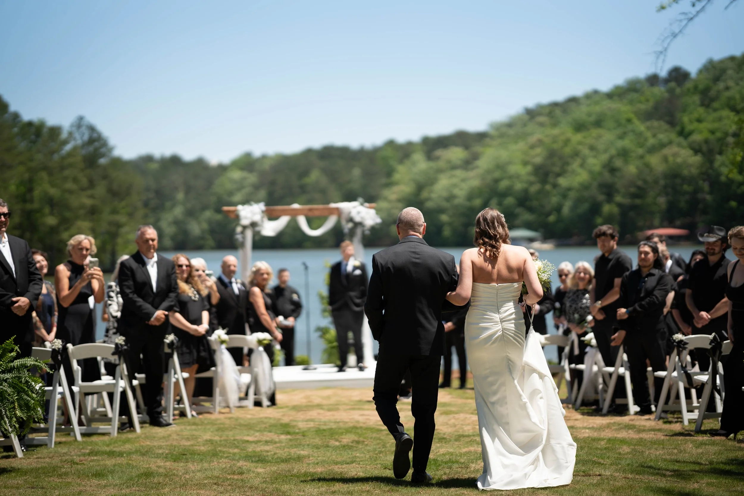 A bride and groom walking towards the wedding ceremony outdoors by a lake with guests seated on white chairs, surrounded by lush green trees and a blue sky.
